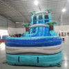 Front view of 20' Bahama Blast Hybrid DL water slide showing vibrant blue colors, inflatable palm trees, and a large splash pool area with wave patterns. The slide features both open and enclosed sliding sections, displayed in an indoor facility with other inflatables visible.