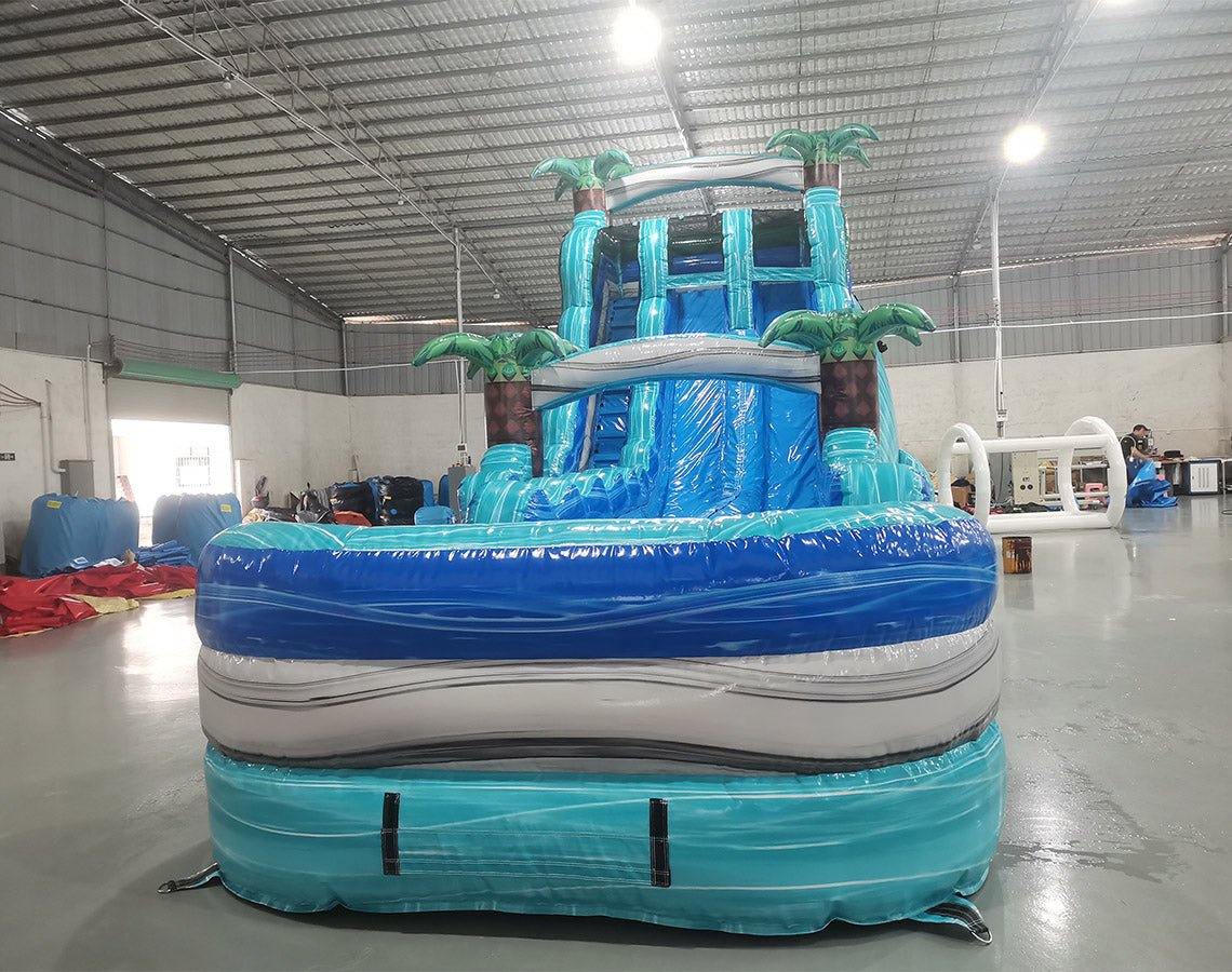 Front view of 20' Bahama Blast Hybrid DL water slide showing vibrant blue colors, inflatable palm trees, and a large splash pool area with wave patterns. The slide features both open and enclosed sliding sections, displayed in an indoor facility with other inflatables visible.