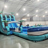 Large inflatable water slide with tropical theme, featuring blue sliding lanes, palm tree decorations, and a spacious splash pool area. The slide combines open and enclosed sections, set up in an indoor warehouse with metal roofing and other equipment visible. Yellow air blowers can be seen at the base of the slide.