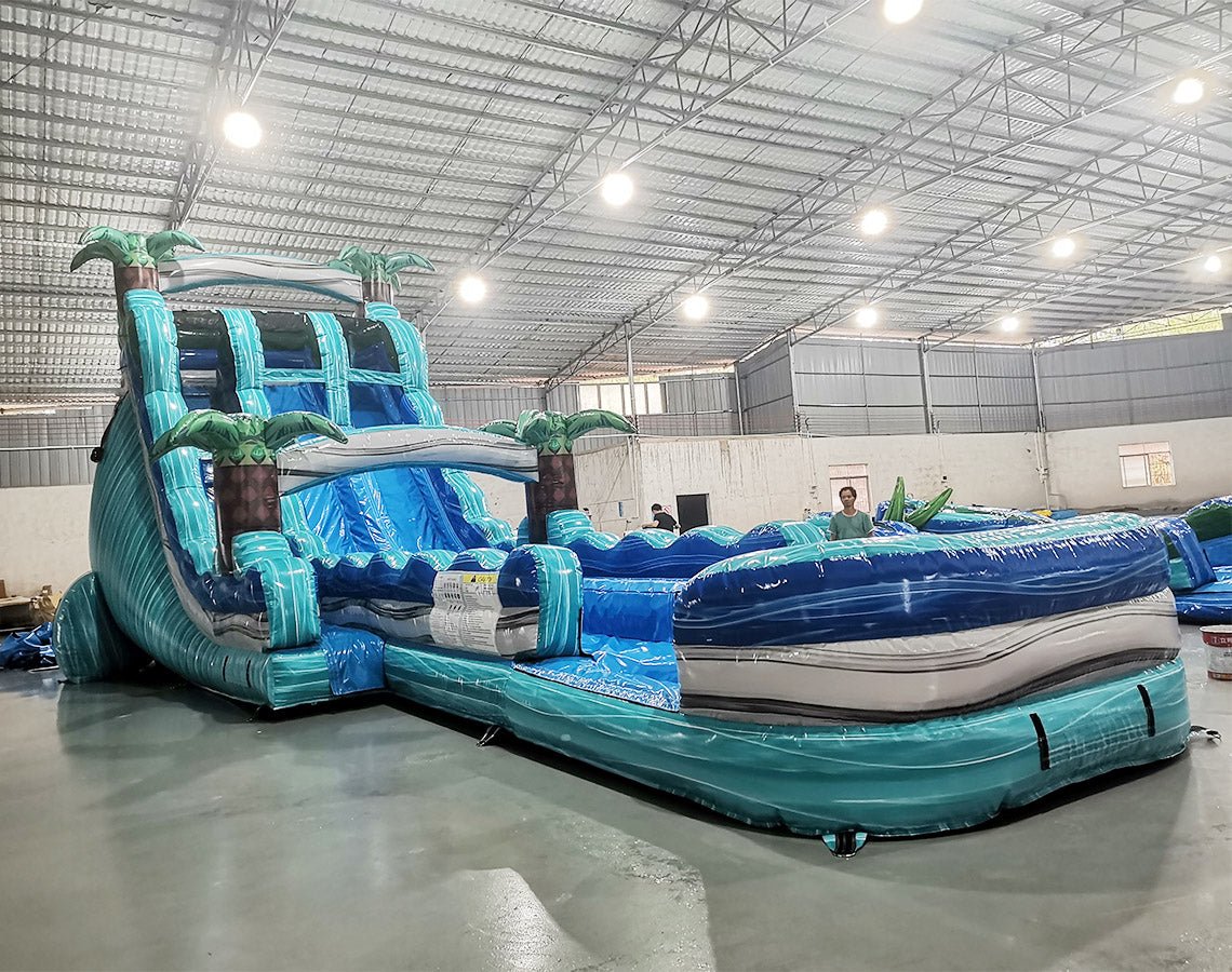 Large inflatable water slide with tropical theme, featuring blue sliding lanes, palm tree decorations, and a spacious splash pool area. The slide combines open and enclosed sections, set up in an indoor warehouse with metal roofing and other equipment visible. Yellow air blowers can be seen at the base of the slide.