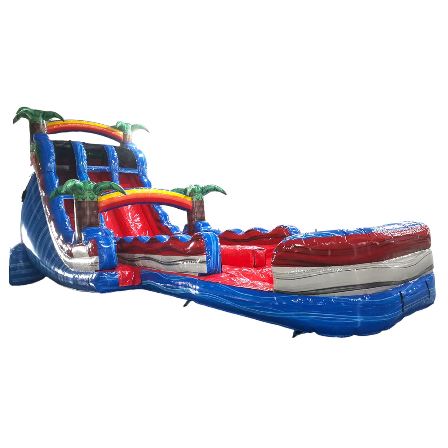 Large inflatable water slide with tropical theme, featuring blue sliding lanes, palm tree decorations, and a spacious splash pool area. The slide combines open and enclosed sections, set up in an indoor warehouse with metal roofing and other equipment visible.