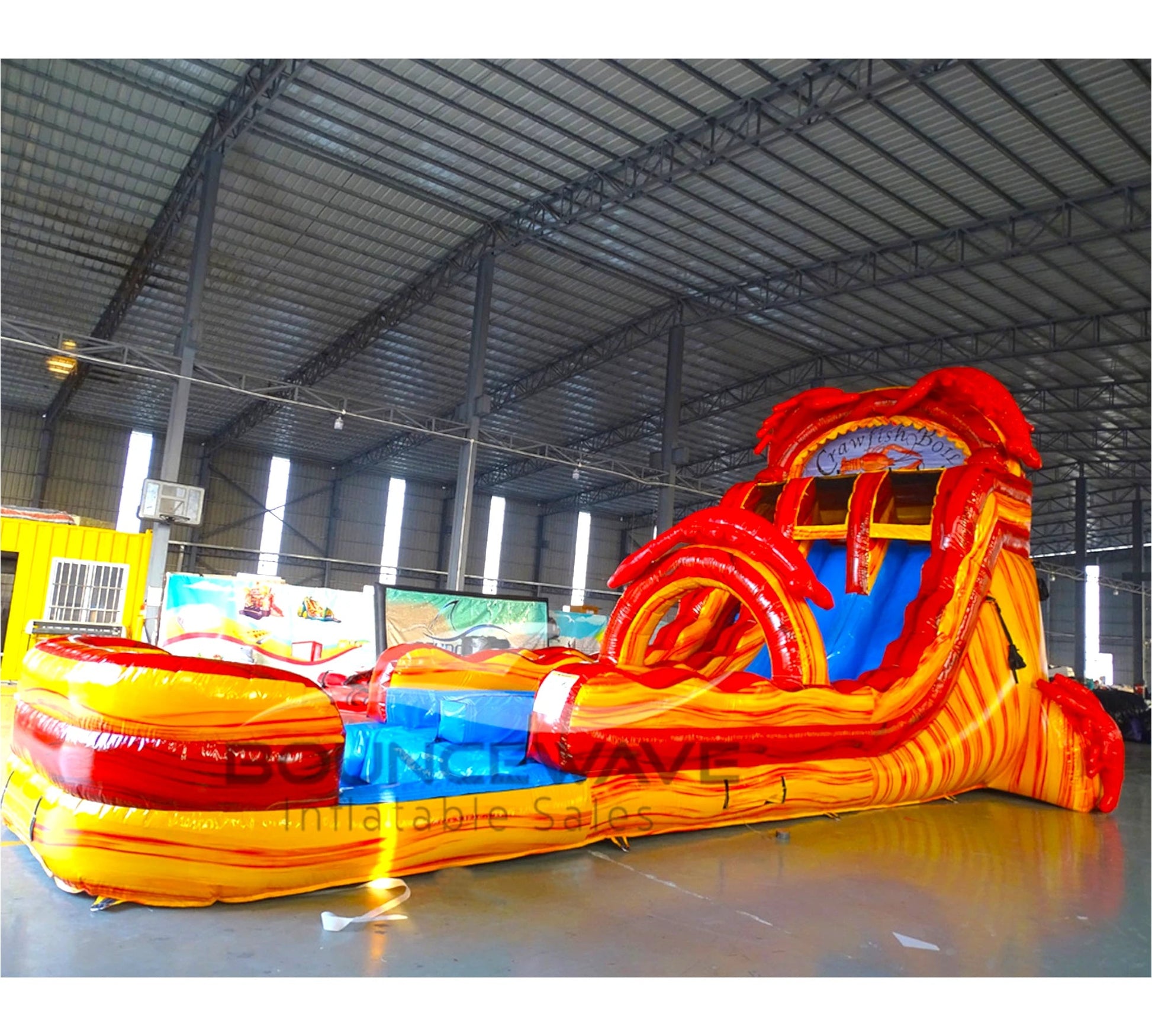 Side view of 20ft Crawfish Hybrid DL water slide showing bright red and yellow design, crawfish-inspired elements, and a large splash pool area. The slide features both open and enclosed sliding sections, displayed in an indoor facility with other inflatables visible.