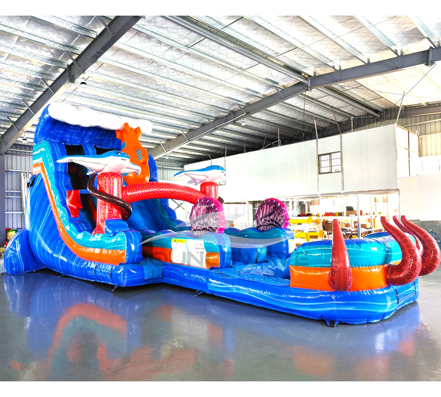 Angled view of 20ft Deep Sea Dive Hybrid DL water slide featuring blue color scheme, shark figures, octopus tentacles, and a large splash pool area. The slide is set up in an indoor warehouse with metal roofing and other equipment visible in the background.