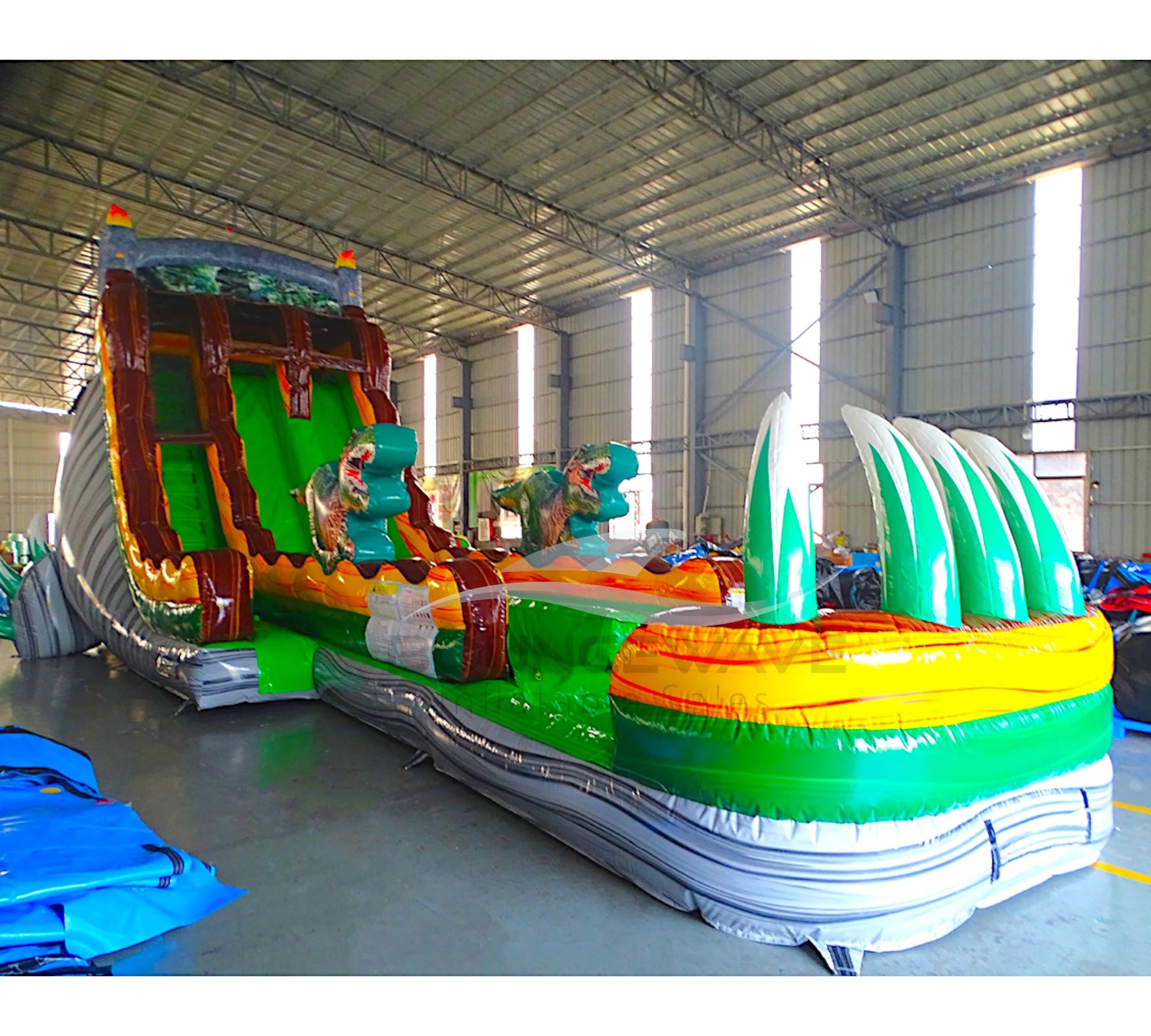 Angled view of 20ft Dino Dive Hybrid DL water slide featuring green color scheme, dinosaur figures, and a large splash pool area. The slide is set up in an indoor warehouse with metal roofing and other equipment visible in the background.