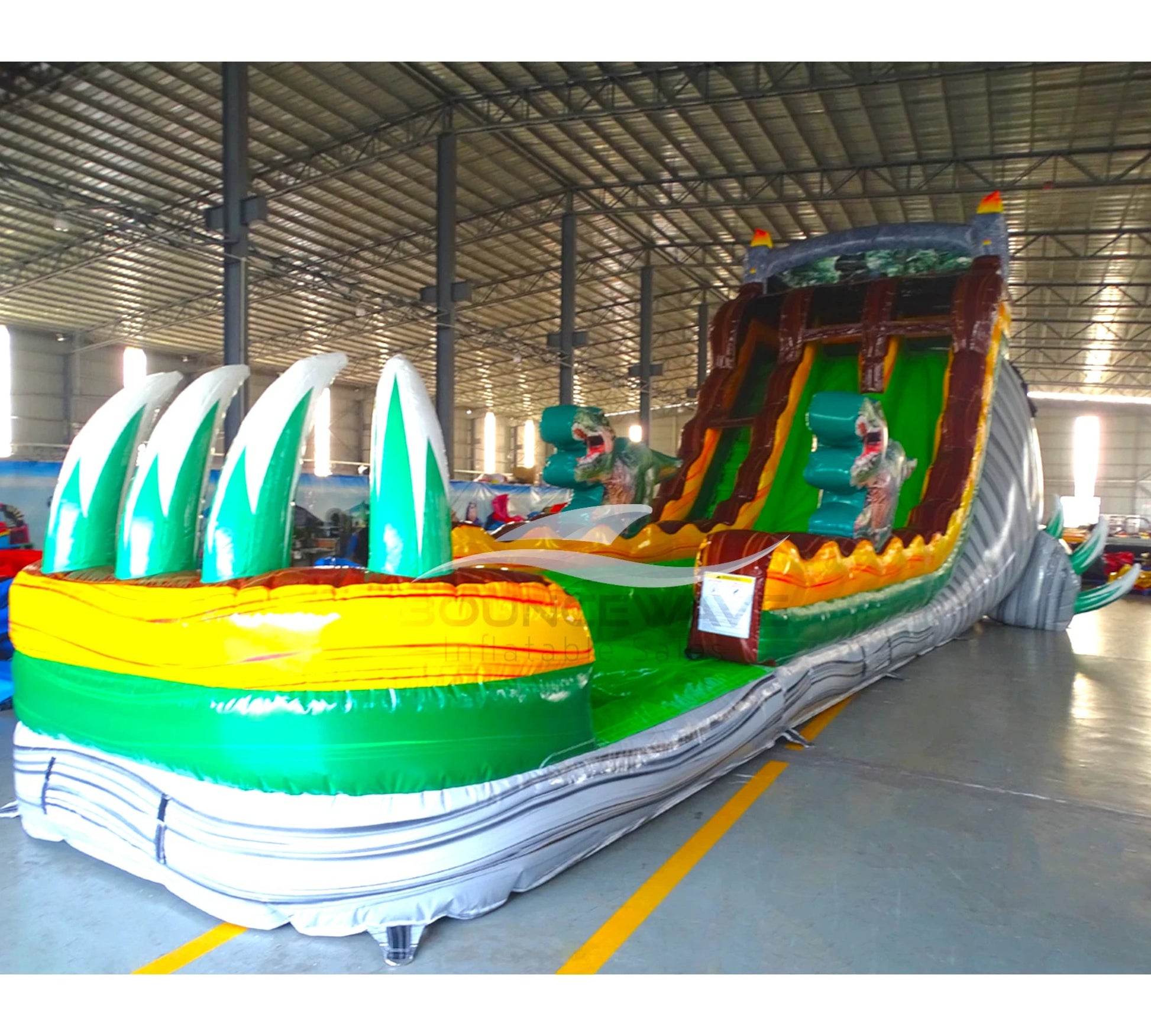 Side view of 20ft Dino Dive Hybrid DL water slide showing vibrant green colors, inflatable dinosaur figures, and a large splash pool area. The slide features both open and enclosed sliding sections, displayed in an indoor facility with other inflatables visible.