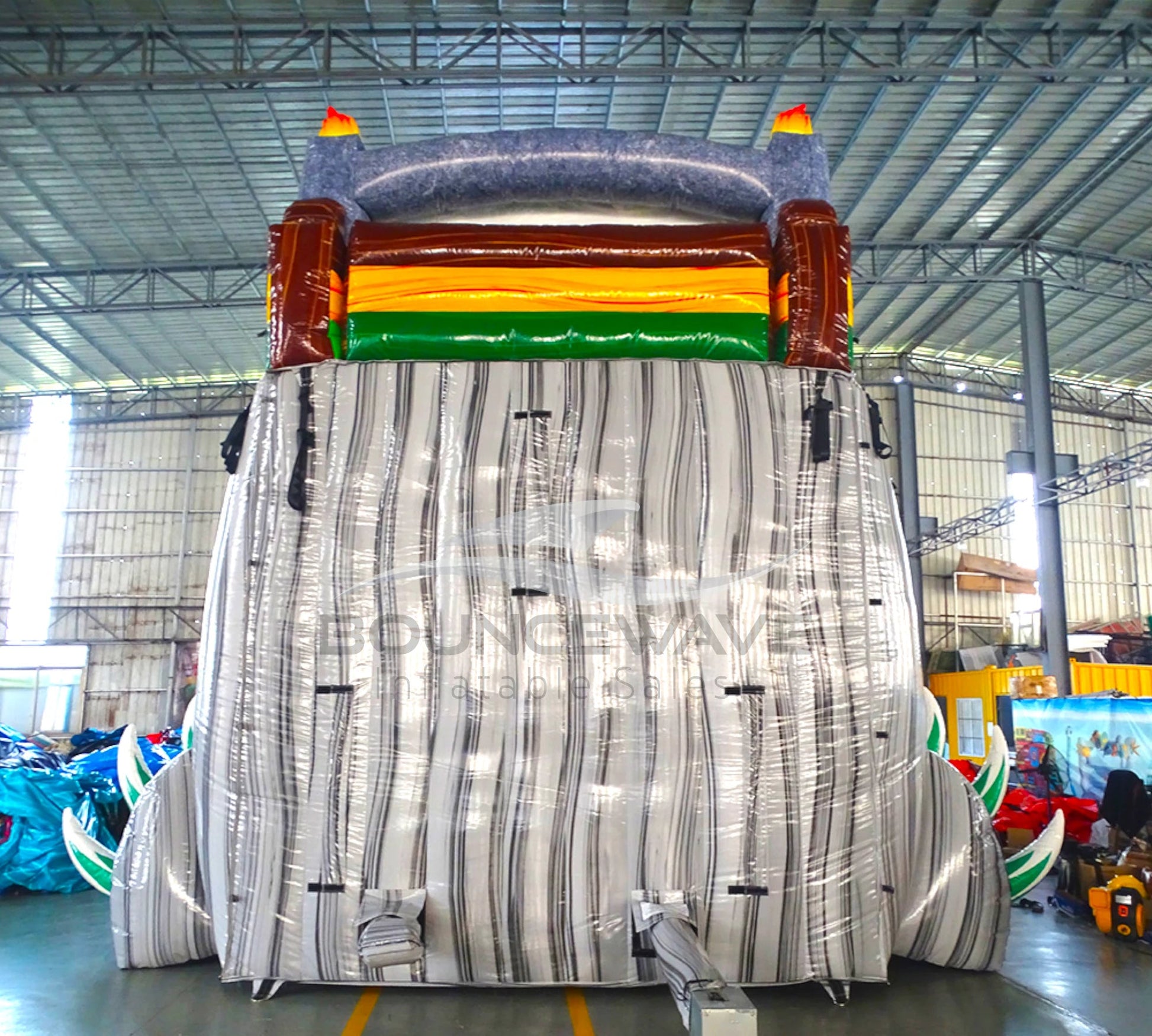 Large inflatable water slide with dinosaur theme, featuring green sliding lanes, T-Rex decorations, and a spacious splash pool area with inflatable dinosaur claws. The slide combines open and enclosed sections, set up in an indoor warehouse with "BOUNCEWAVE Inflatable Sales" branding visible on the splash pool.