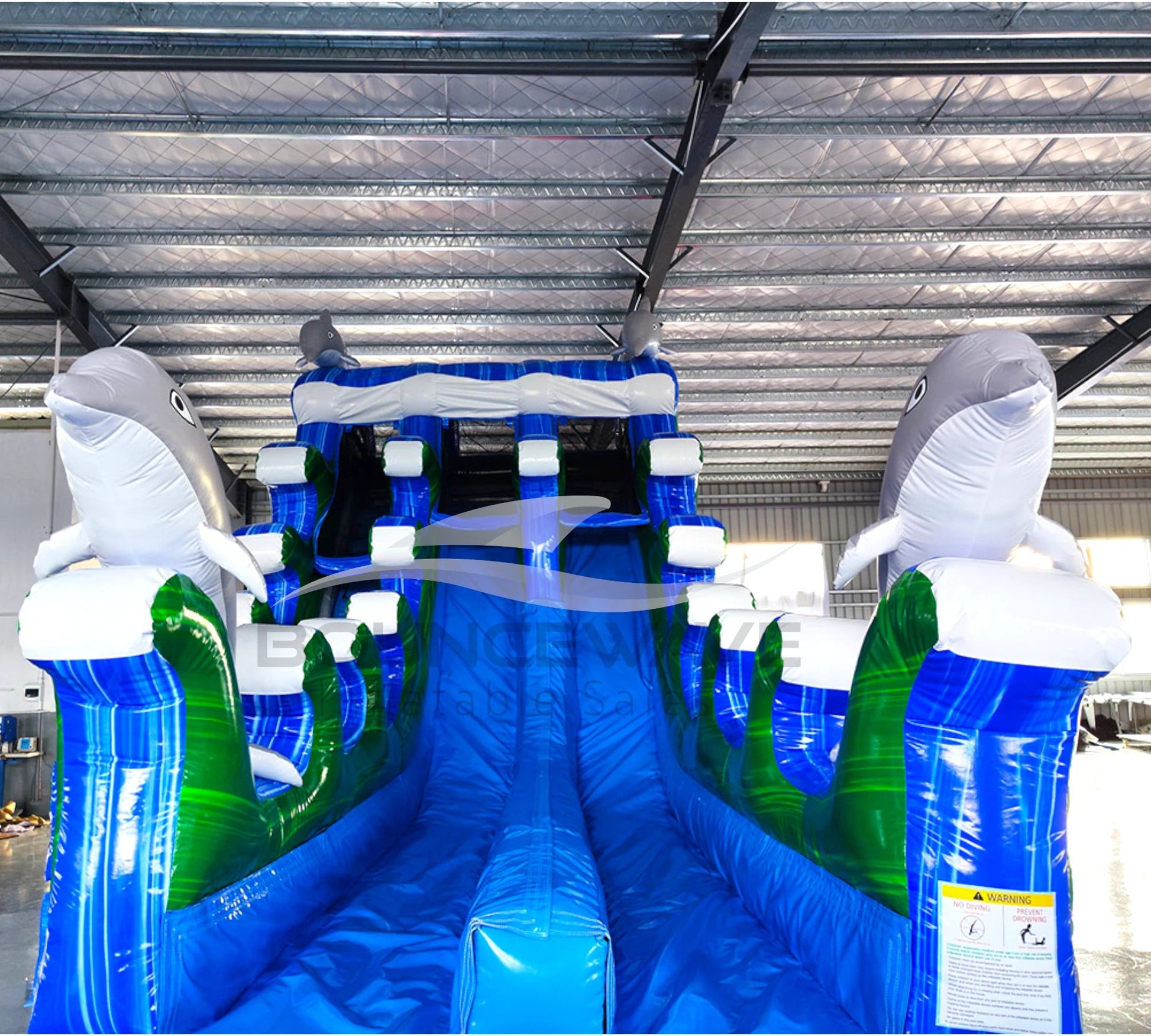 Large inflatable water slide with ocean theme, featuring blue sliding lanes, dolphin decorations, and a spacious splash pool area. The slide combines open and enclosed sections, set up in an indoor warehouse with metal roofing visible.