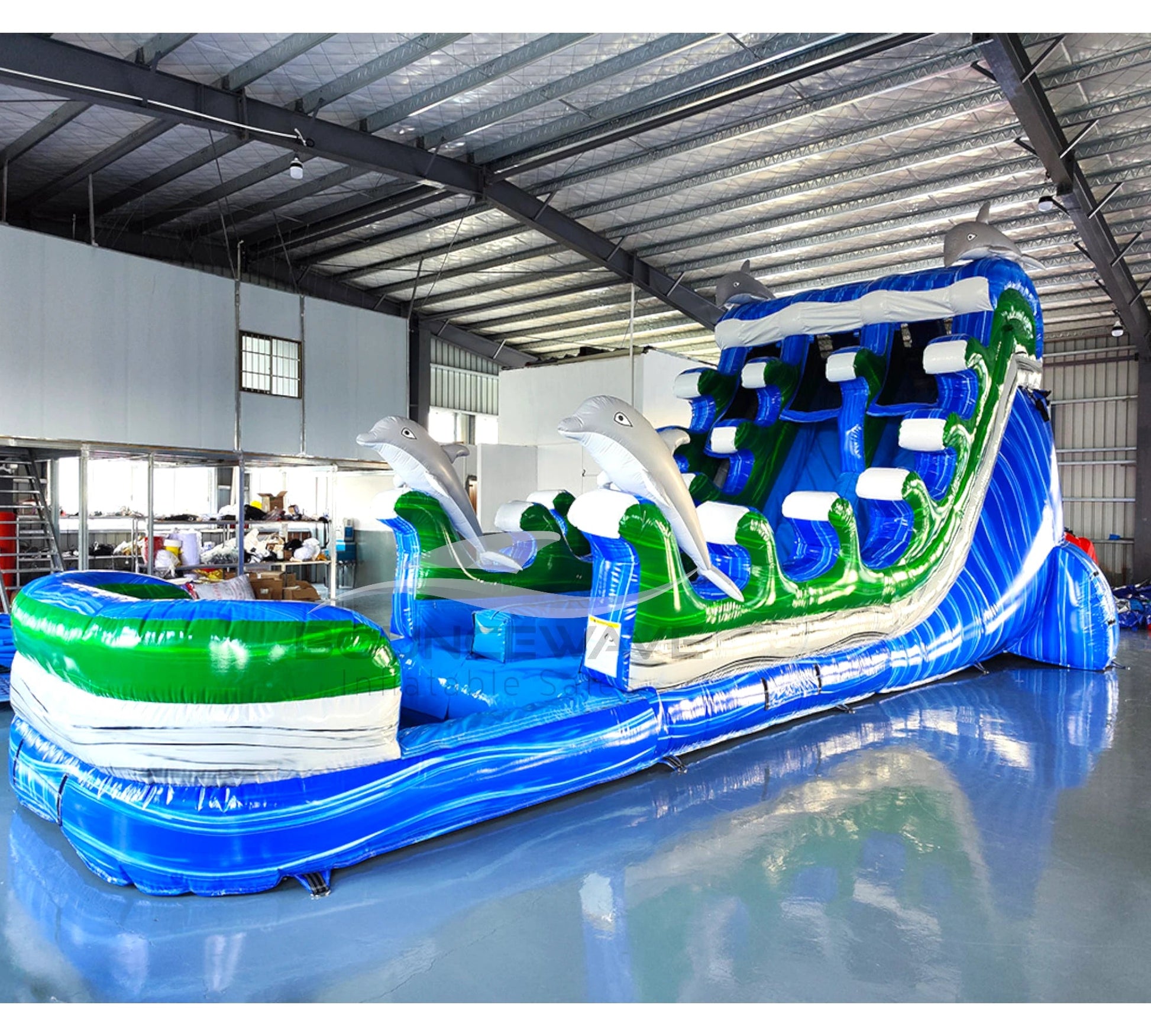 Large inflatable water slide with ocean theme, featuring blue sliding lanes, dolphin decorations, and a spacious splash pool area. The slide combines open and enclosed sections, set up in an indoor warehouse with metal roofing visible.