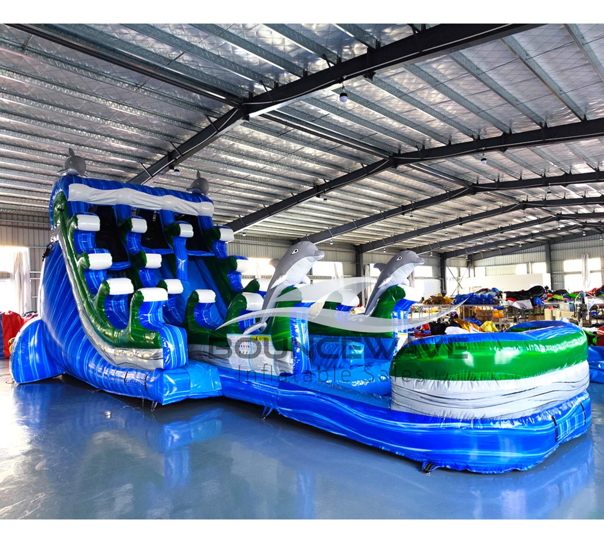 Angled view of 20ft Dolphin Dive Hybrid DL water slide featuring blue color scheme, dolphin figures, and a large splash pool area. The slide is set up in an indoor warehouse with metal roofing and other equipment visible in the background.