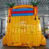 Large inflatable water slide with tropical theme, featuring vibrant red, yellow, and blue colors, palm tree decorations, and a spacious splash pool area. The slide combines open and enclosed sections, set up in an indoor warehouse with metal roofing and other equipment visible.