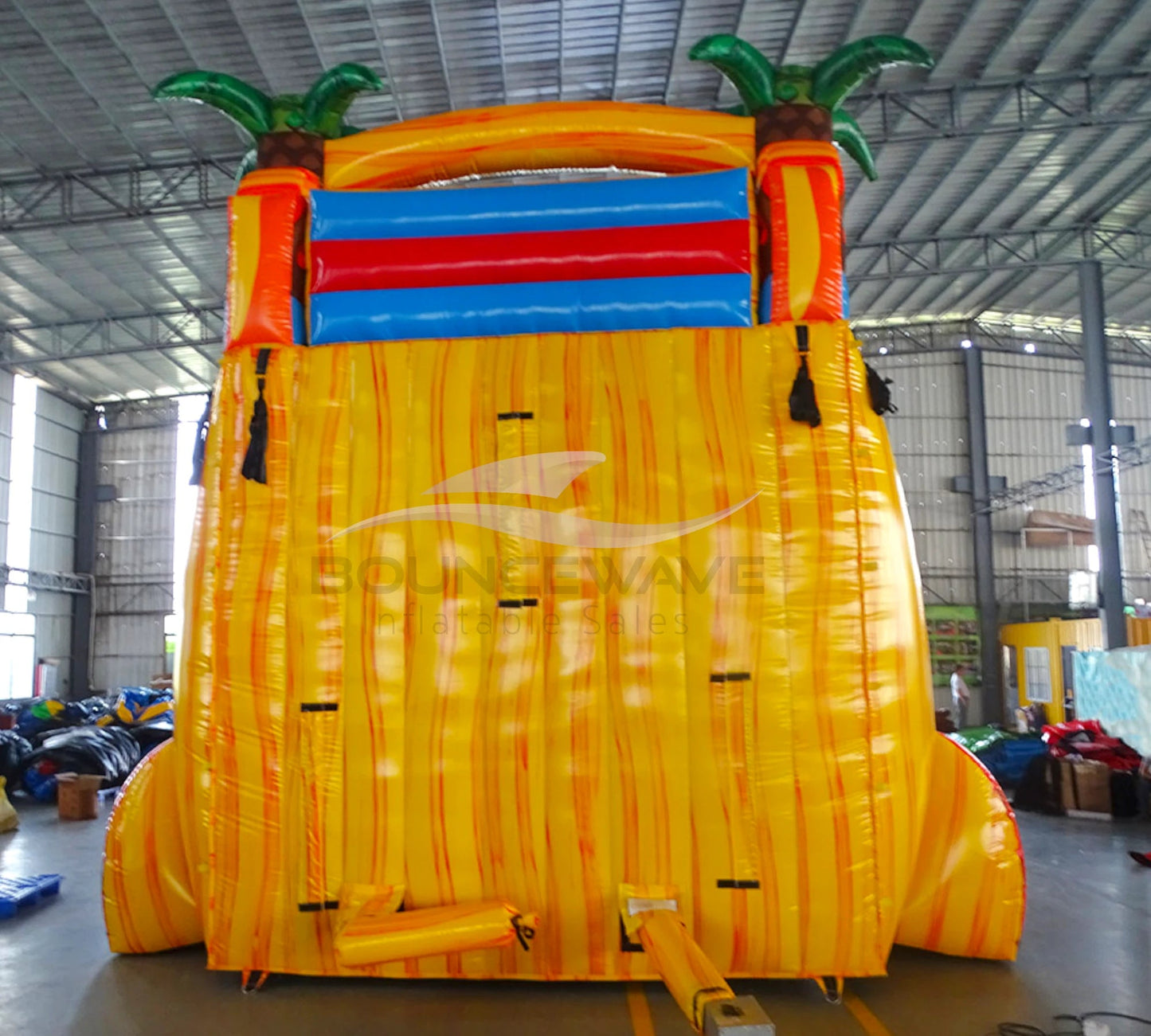 Large inflatable water slide with tropical theme, featuring vibrant red, yellow, and blue colors, palm tree decorations, and a spacious splash pool area. The slide combines open and enclosed sections, set up in an indoor warehouse with metal roofing and other equipment visible.