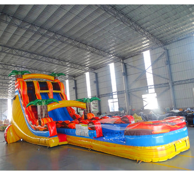 Rear view of 20ft Fiesta Fire Hybrid DL water slide featuring red and yellow colors, inflatable palm trees, and enclosed tube slide sections. The slide is set up in an indoor facility with metal roofing and other inflatables visible in the background.