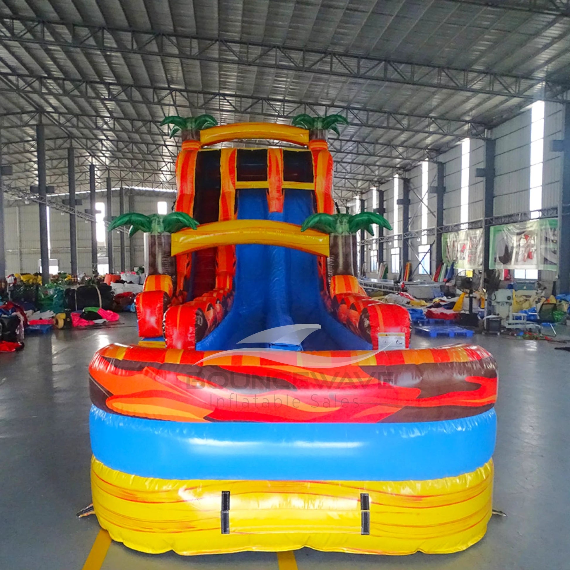 Front view of 20ft Fiesta Fire Hybrid DL water slide showing bright red and yellow design, inflatable palm trees, and a large splash pool area with "BOUNCEWAVE Inflatable Sales" branding. The slide features both open and enclosed sliding sections.