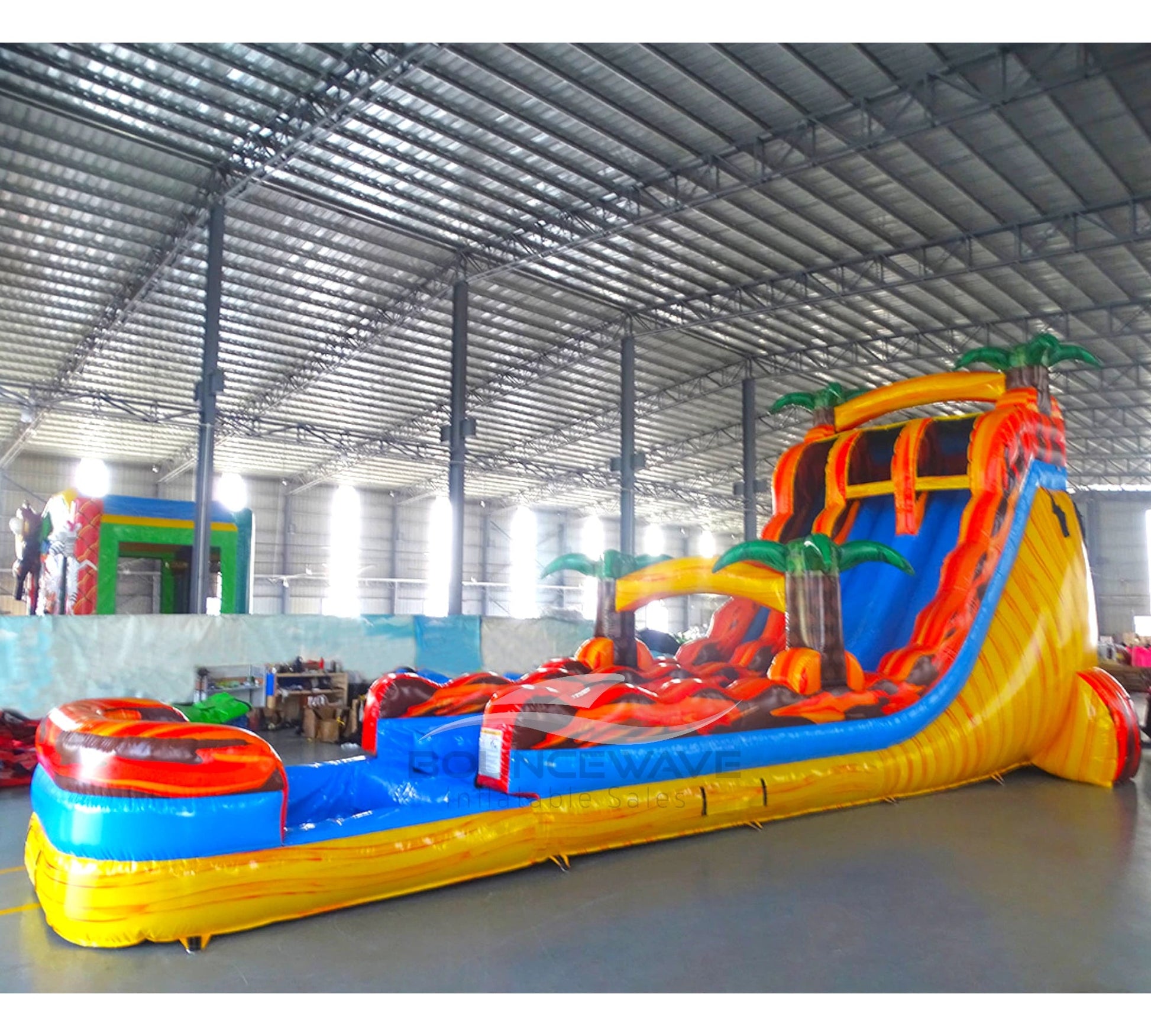 Side angle view of the 20ft Fiesta Fire Hybrid DL inflatable water slide, showcasing its red and yellow color scheme, palm tree decorations, and spacious splash pool. The slide combines open and tube slide elements, set up in an indoor warehouse with other equipment visible.