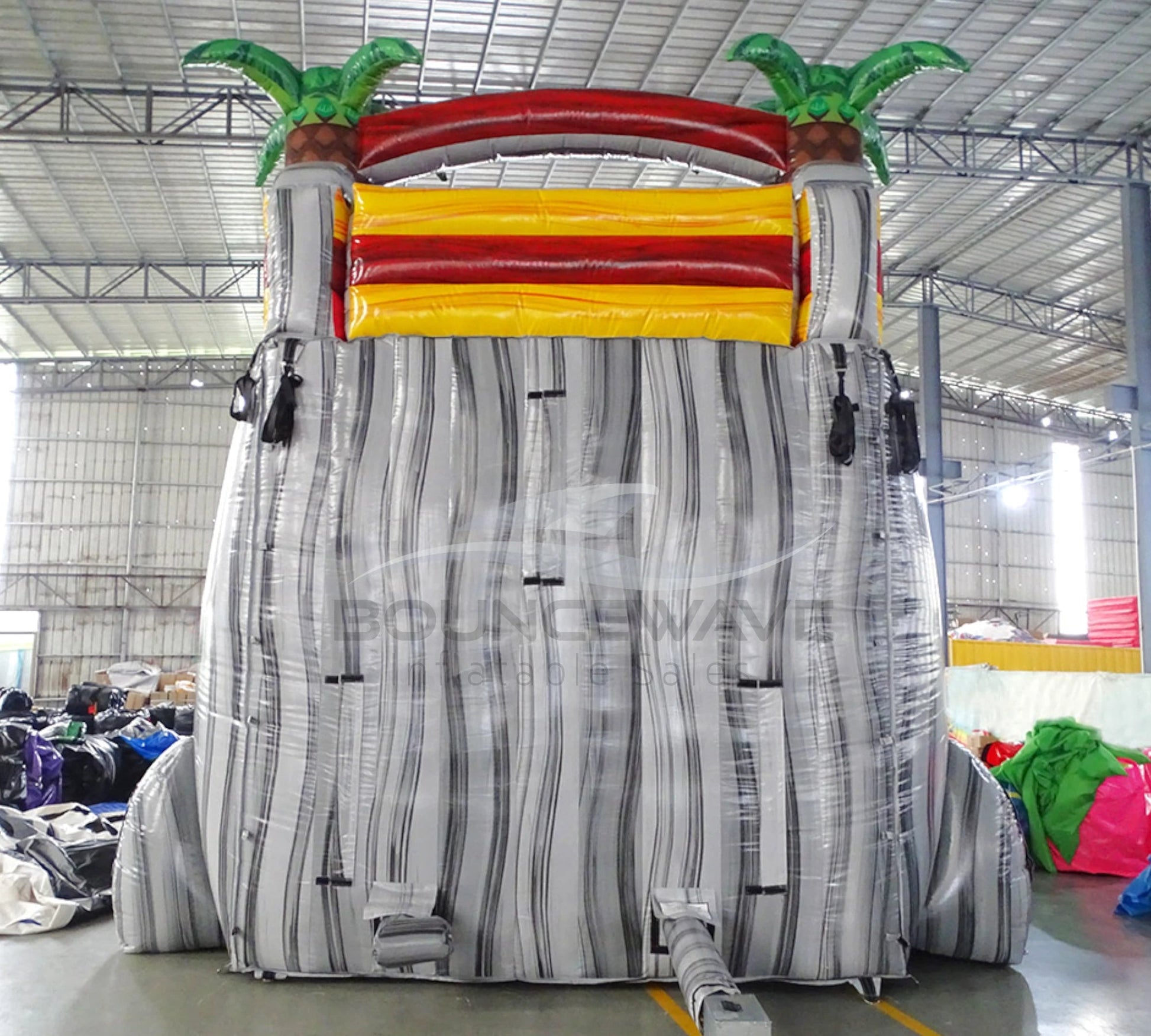 Large inflatable water slide with tropical theme, featuring vibrant red, yellow, and white colors, palm tree decorations, and a spacious splash pool area. The slide combines open and enclosed sections, set up in an indoor warehouse with metal roofing and other equipment visible in the background.