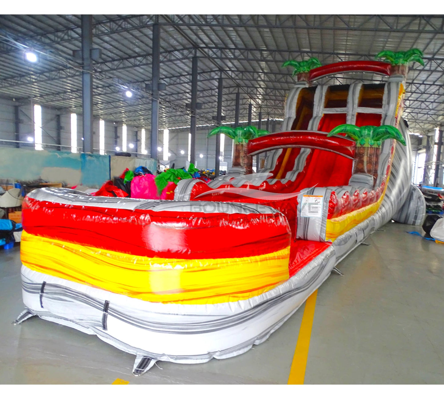 Large inflatable water slide with tropical theme, featuring vibrant red, yellow, and white colors, palm tree decorations, and a spacious splash pool area. The slide combines open and enclosed sections, set up in an indoor warehouse with metal roofing visible.