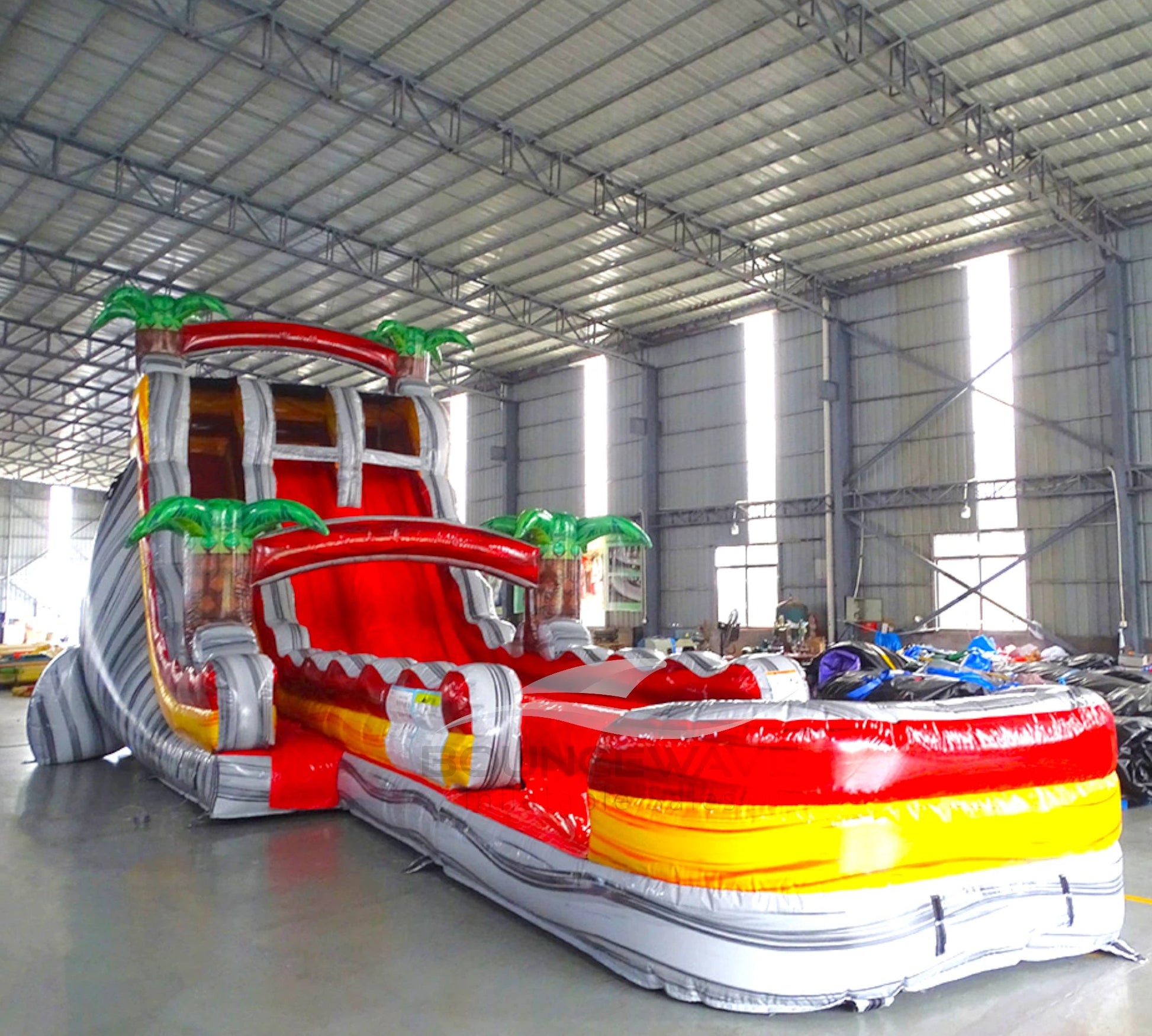 Front angle view of the 20ft Fire Island Hybrid DL inflatable water slide, showcasing its red, yellow, and white color scheme, palm tree decorations, and spacious splash pool. The slide combines open and tube slide elements, set up in an indoor warehouse with other inflatables visible.