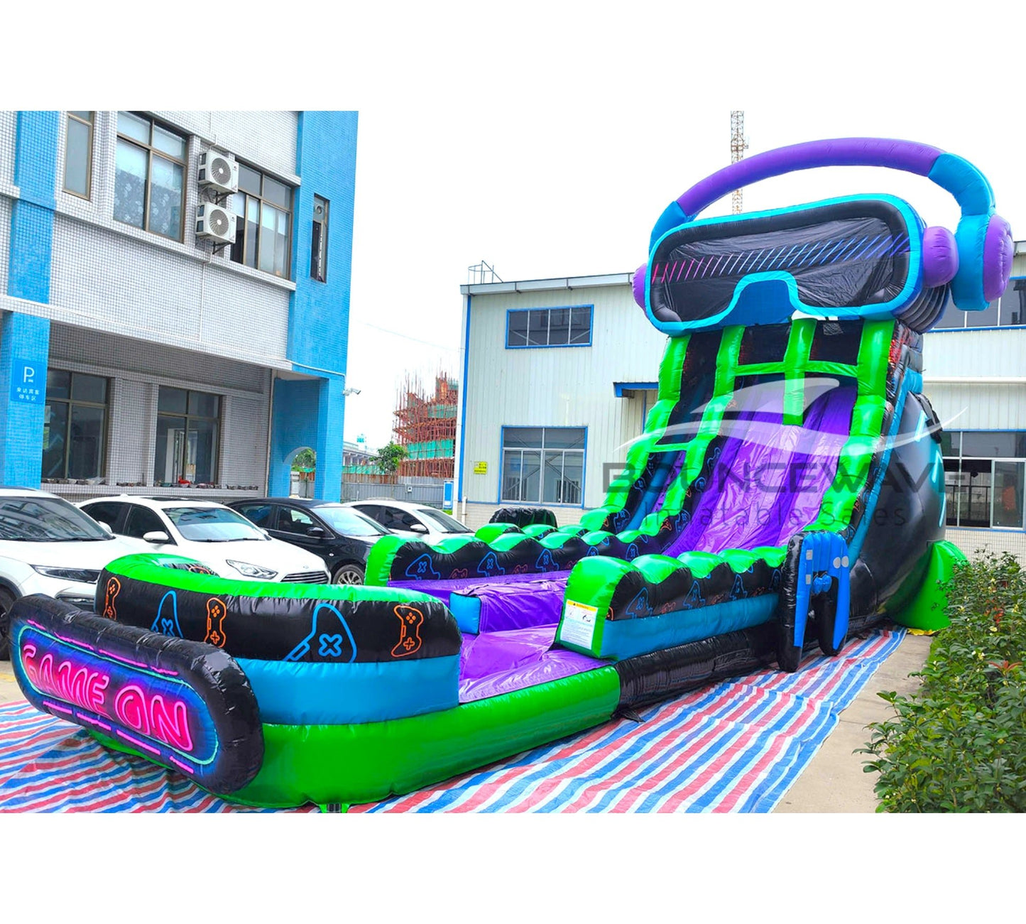 Side view of 20ft GameWave Hybrid DL water slide showing vibrant green, purple, and blue colors, giant goggles and headphones design, and a large splash pool area with "GAME ON" neon sign. The slide is set up in a parking lot with buildings visible.