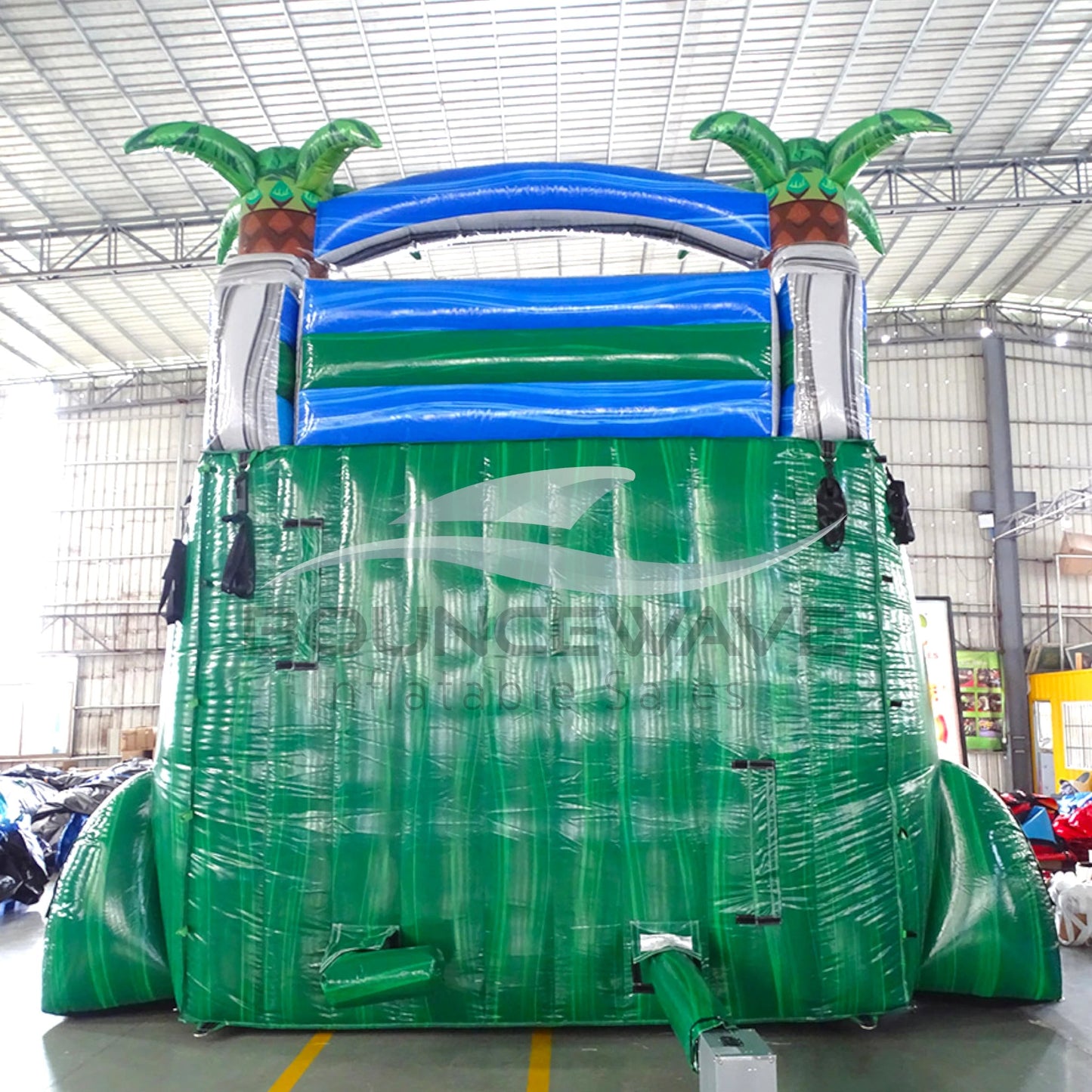 Large inflatable water slide with tropical theme, featuring blue sliding lanes, palm tree decorations, and a spacious splash pool area. The slide combines open and enclosed sections, set up in an indoor warehouse with metal roofing and other equipment visible in the background.