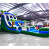 Large inflatable water slide with tropical theme, featuring blue sliding lanes, palm tree decorations, and a spacious splash pool area. The slide combines open and enclosed sections, set up in an indoor warehouse with metal roofing visible.