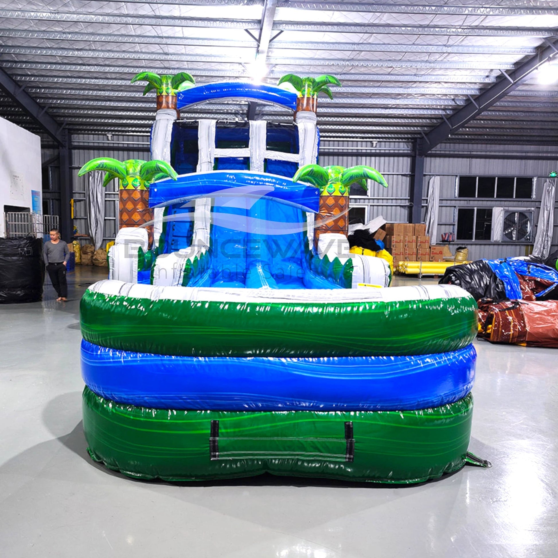 Front view of the 20ft Green Gush Hybrid DL inflatable water slide, showcasing its blue sliding lanes, palm tree decorations, and spacious splash pool with "BOUNCEWAVE" branding. The slide combines open and tube slide elements, set up in an indoor warehouse.