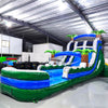 Side view of 20ft Green Gush Hybrid DL water slide showing vibrant blue and green colors, inflatable palm trees, and a large splash pool area. The slide features both open and enclosed sliding sections, displayed in an indoor facility with other inflatables visible.