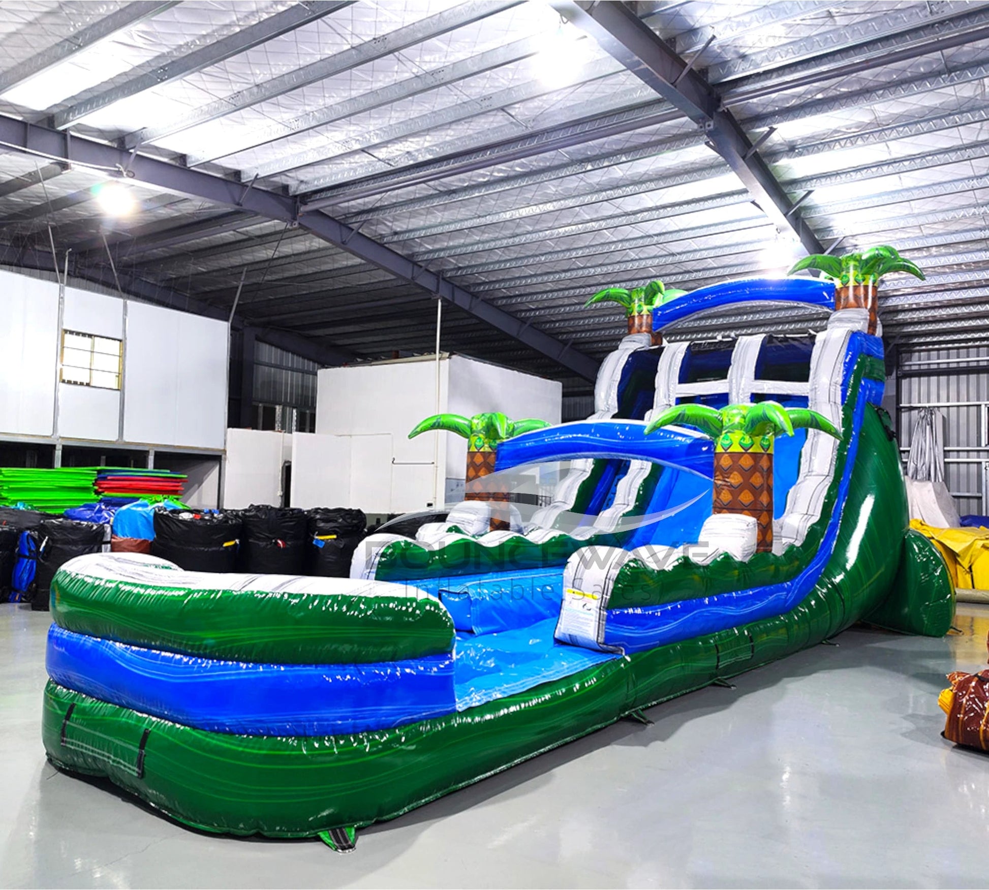 Side view of 20ft Green Gush Hybrid DL water slide showing vibrant blue and green colors, inflatable palm trees, and a large splash pool area. The slide features both open and enclosed sliding sections, displayed in an indoor facility with other inflatables visible.