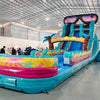 Front angle view of the 20ft Island Flow Hybrid DL inflatable water slide, showcasing its turquoise sliding lanes, palm tree decorations, and spacious splash pool with tropical patterns. The slide is topped with large inflatable sunglasses and is set up in an indoor warehouse with workers visible.