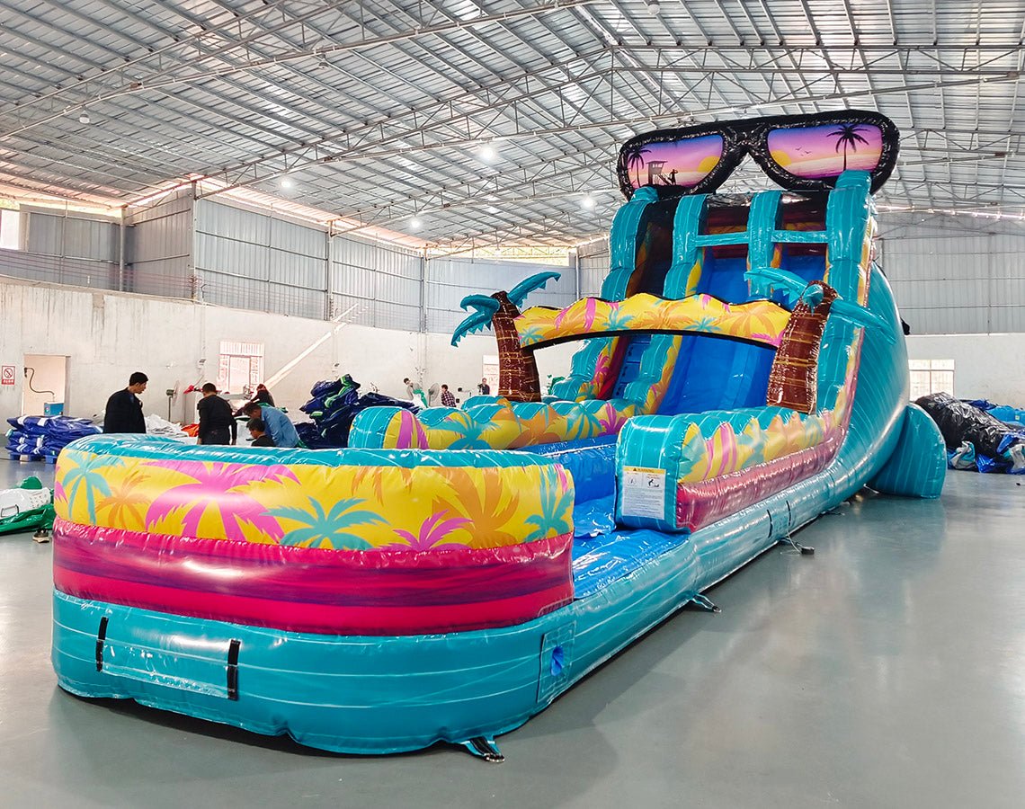 Front angle view of the 20ft Island Flow Hybrid DL inflatable water slide, showcasing its turquoise sliding lanes, palm tree decorations, and spacious splash pool with tropical patterns. The slide is topped with large inflatable sunglasses and is set up in an indoor warehouse with workers visible.