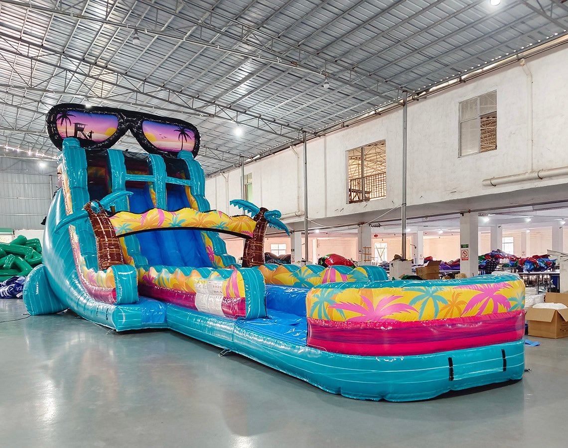 Side view of 20ft Island Flow Hybrid DL water slide showing vibrant turquoise colors, inflatable palm trees, and a large splash pool area with tropical designs. The slide features a giant sunglasses decoration on top and is displayed in an indoor facility with other inflatables visible.