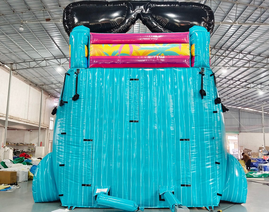 Rear view of 20ft Island Flow Hybrid DL water slide featuring turquoise color scheme, giant inflatable sunglasses on top, and tropical-themed splash pool area. The slide is set up in an indoor facility with metal roofing and other equipment visible in the background.