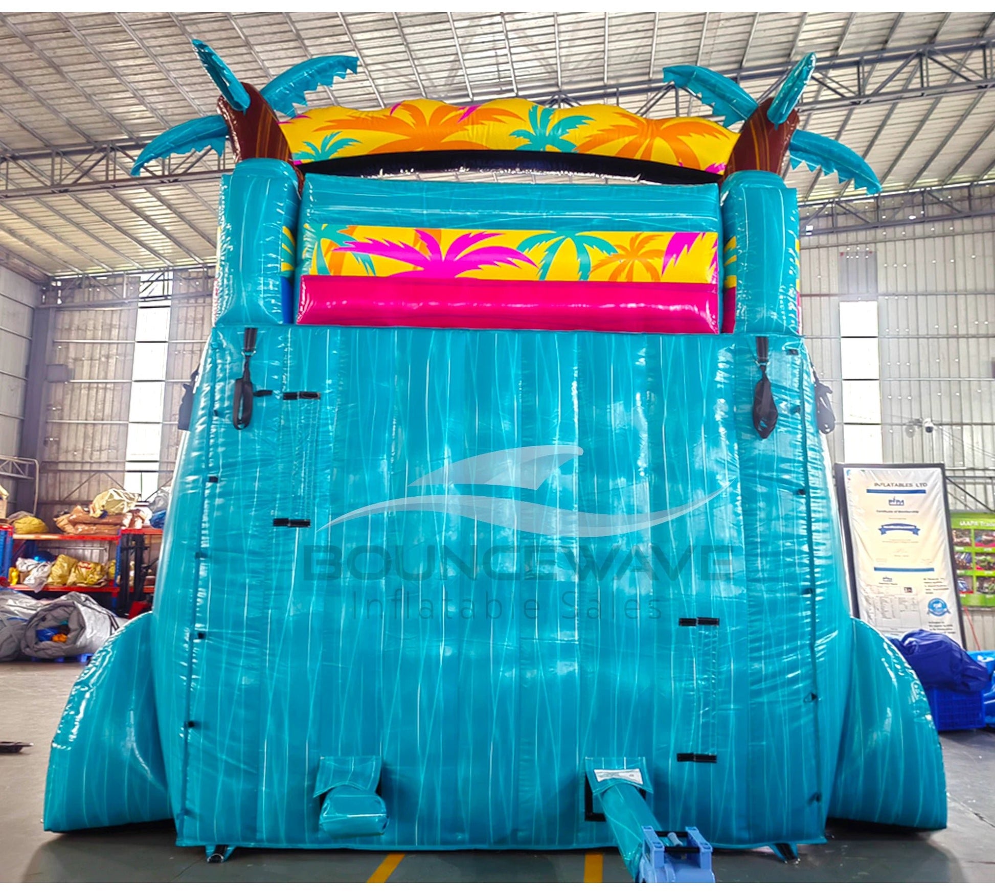 Large inflatable water slide with tropical theme, featuring turquoise sliding lanes, palm tree decorations, and a spacious splash pool area with colorful palm leaf designs. The slide combines open and enclosed sections, set up in an indoor warehouse with metal roofing and other equipment visible in the background.