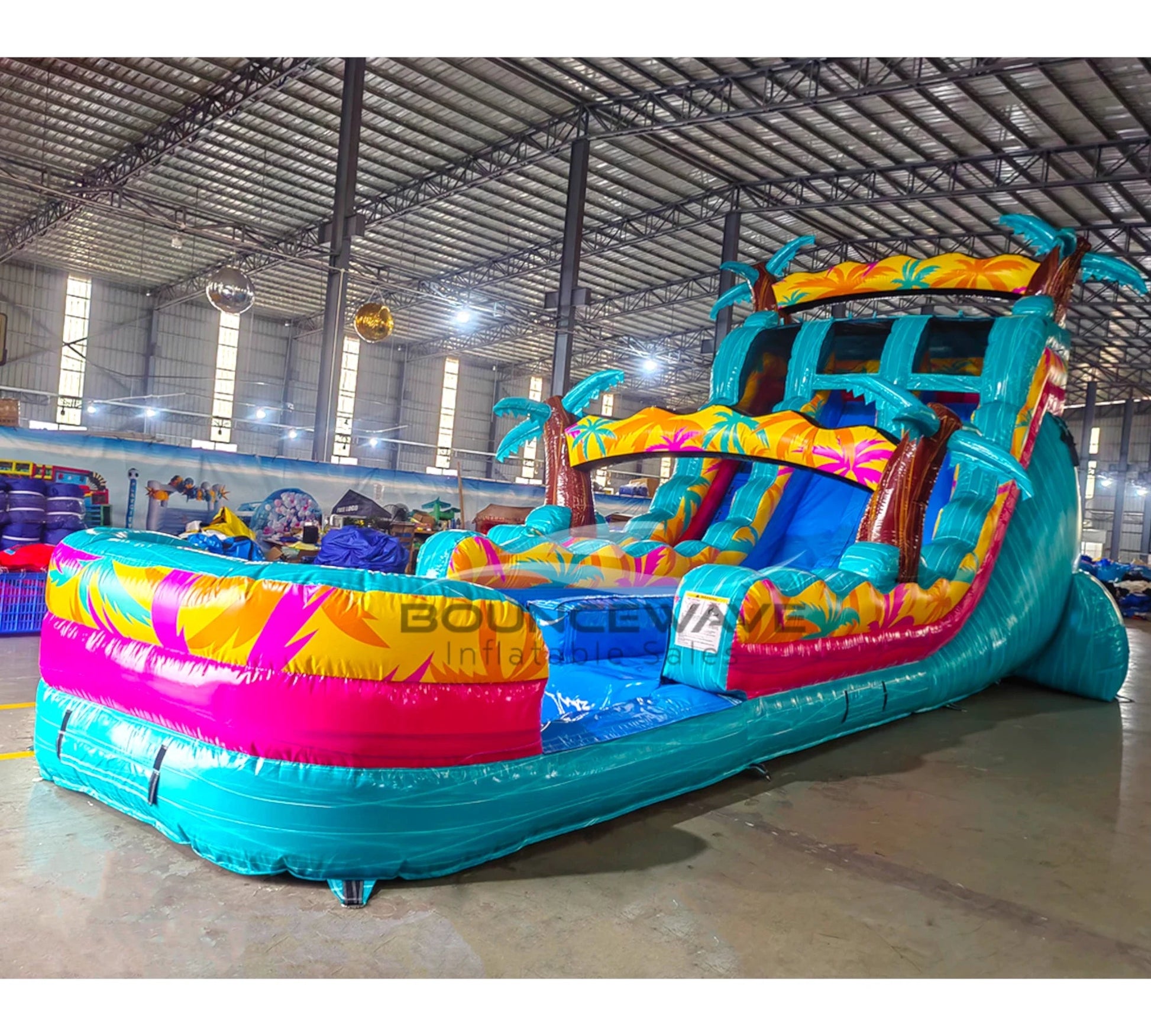 Angled view of 20ft Island Flow Palms Hybrid DL water slide featuring turquoise color scheme, palm tree decorations, and a large splash pool area with vibrant leaf patterns. The slide is set up in an indoor facility with metal roofing and other inflatables visible in the background.