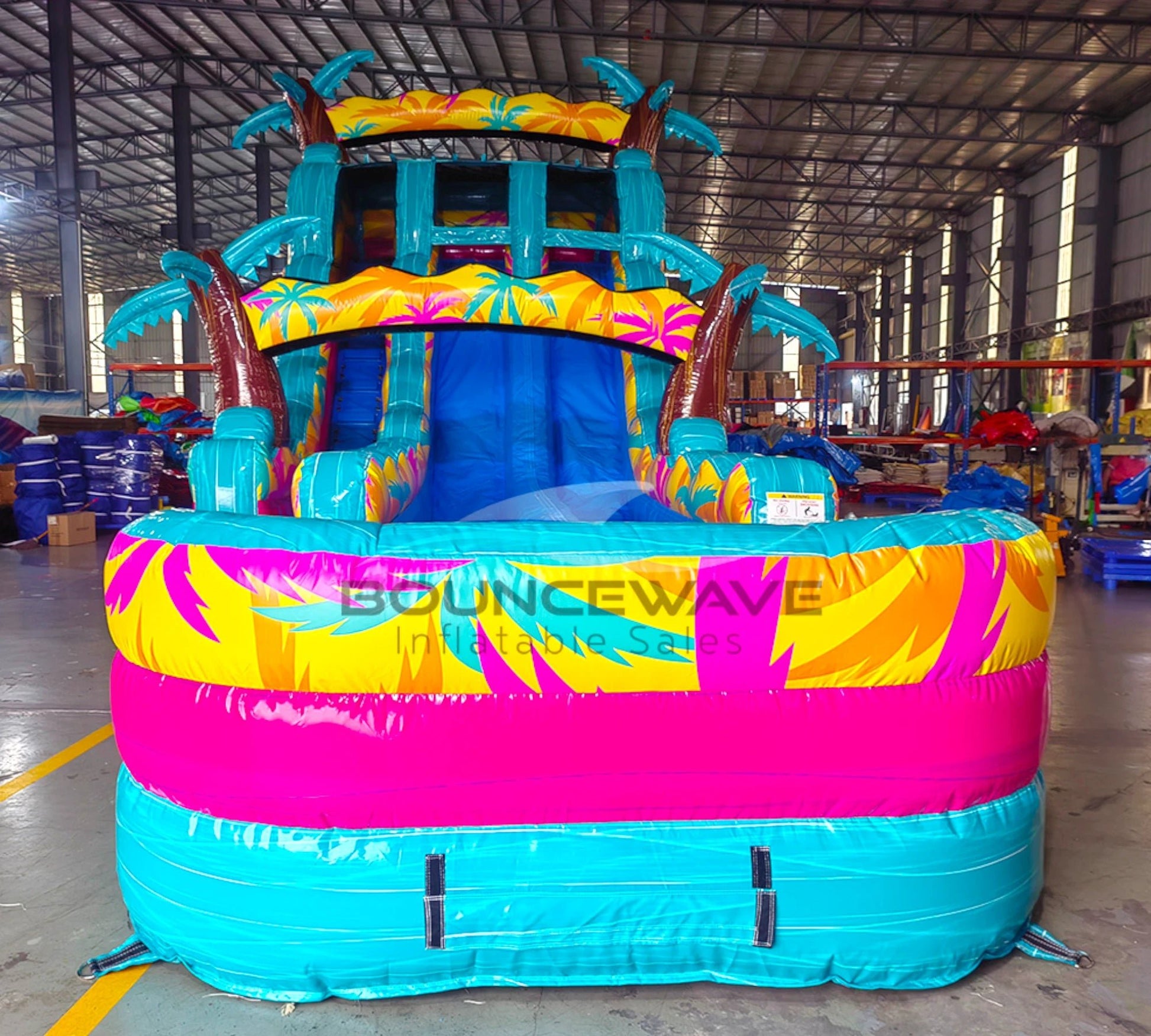 Large inflatable water slide with tropical theme, featuring turquoise sliding lanes, palm tree decorations, and a spacious splash pool area with colorful palm leaf designs. The slide has a "BOUNCEWAVE Inflatable Sales" logo and is set up in an indoor warehouse with metal roofing visible.