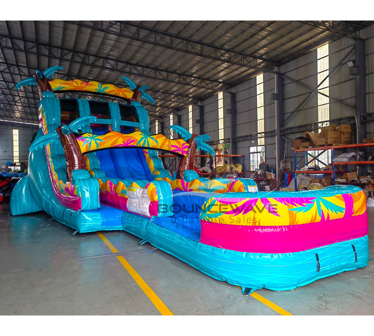 Front angle view of the 20ft Island Flow Palms Hybrid DL inflatable water slide, showcasing its turquoise sliding lanes, palm tree decorations, and spacious splash pool with colorful tropical designs. The slide is set up in an indoor warehouse with other equipment and inflatables visible.