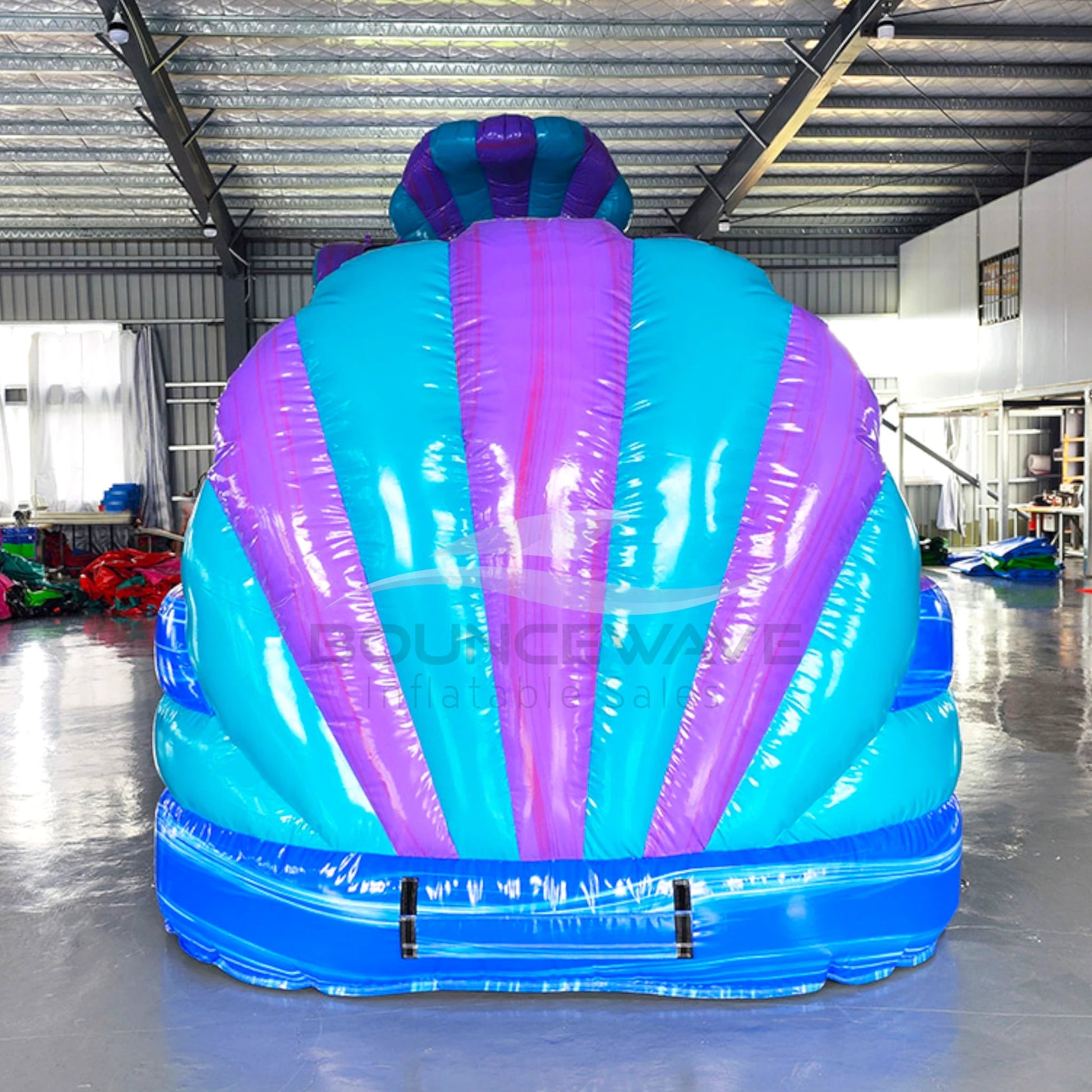 Large inflatable water slide with ocean theme, featuring blue and purple colors, shell-shaped top, and mermaid decorations. The slide has both open and enclosed sections, a spacious splash pool area, and is set up in an indoor warehouse with metal roofing visible.