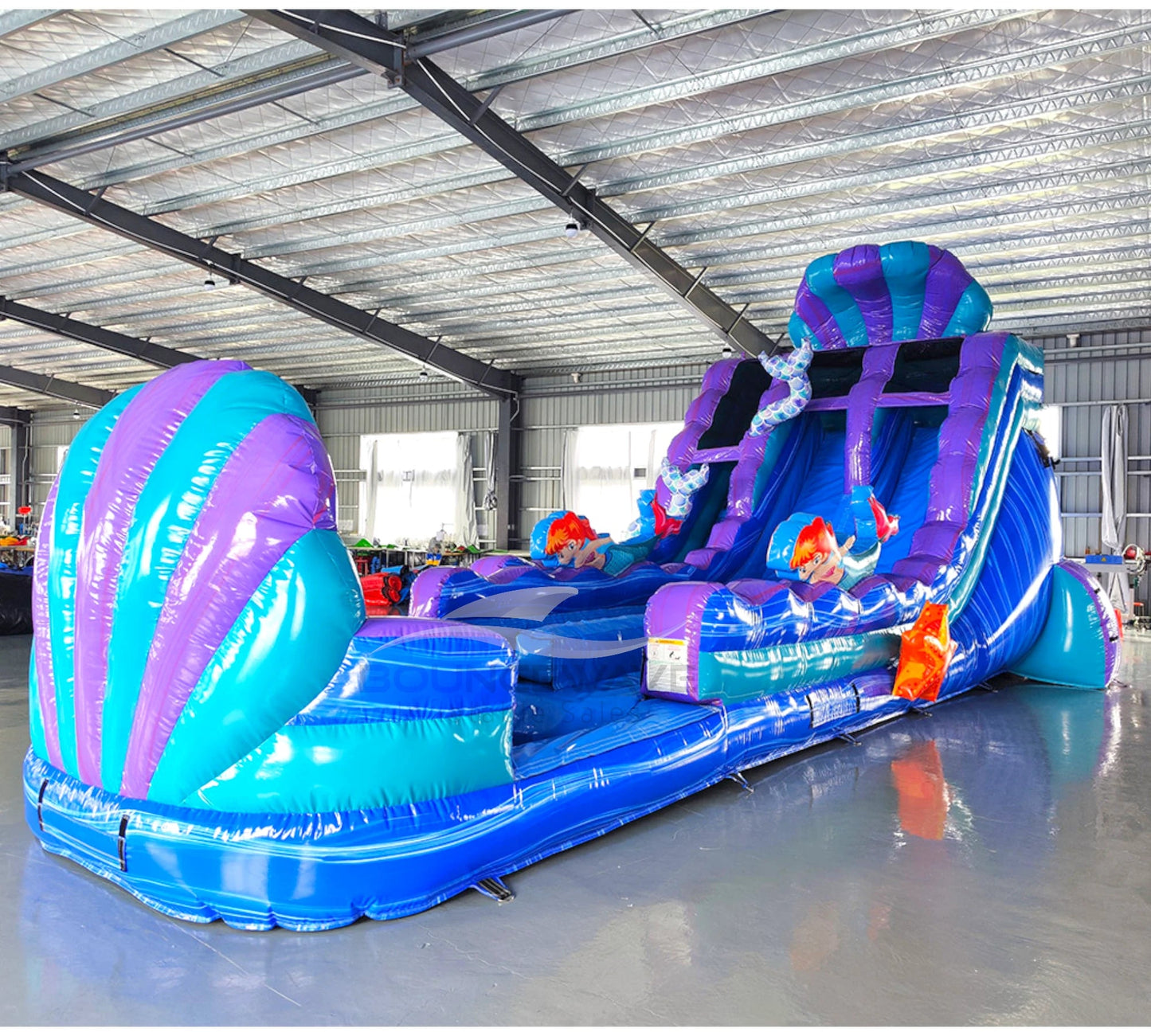 Angled view of 20ft Mermaid's Cove Hybrid DL water slide featuring blue, purple, and turquoise color scheme, shell-shaped design, mermaid figures, and a large splash pool area. The slide is set up in an indoor facility with metal roofing and other inflatables visible in the background.