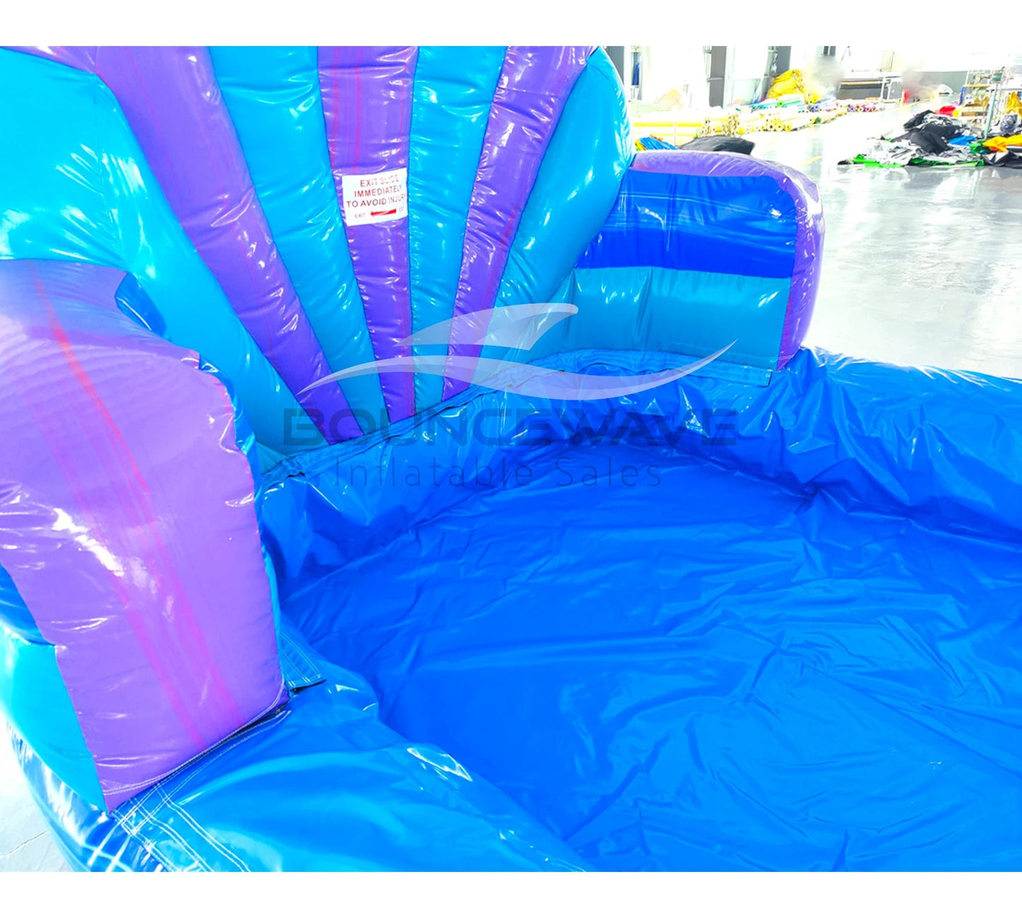 Large inflatable water slide with ocean theme, featuring blue and purple colors, shell-shaped top, and mermaid decorations. The slide has both open and enclosed sections, a spacious splash pool area, and is set up in an indoor warehouse with metal roofing visible.