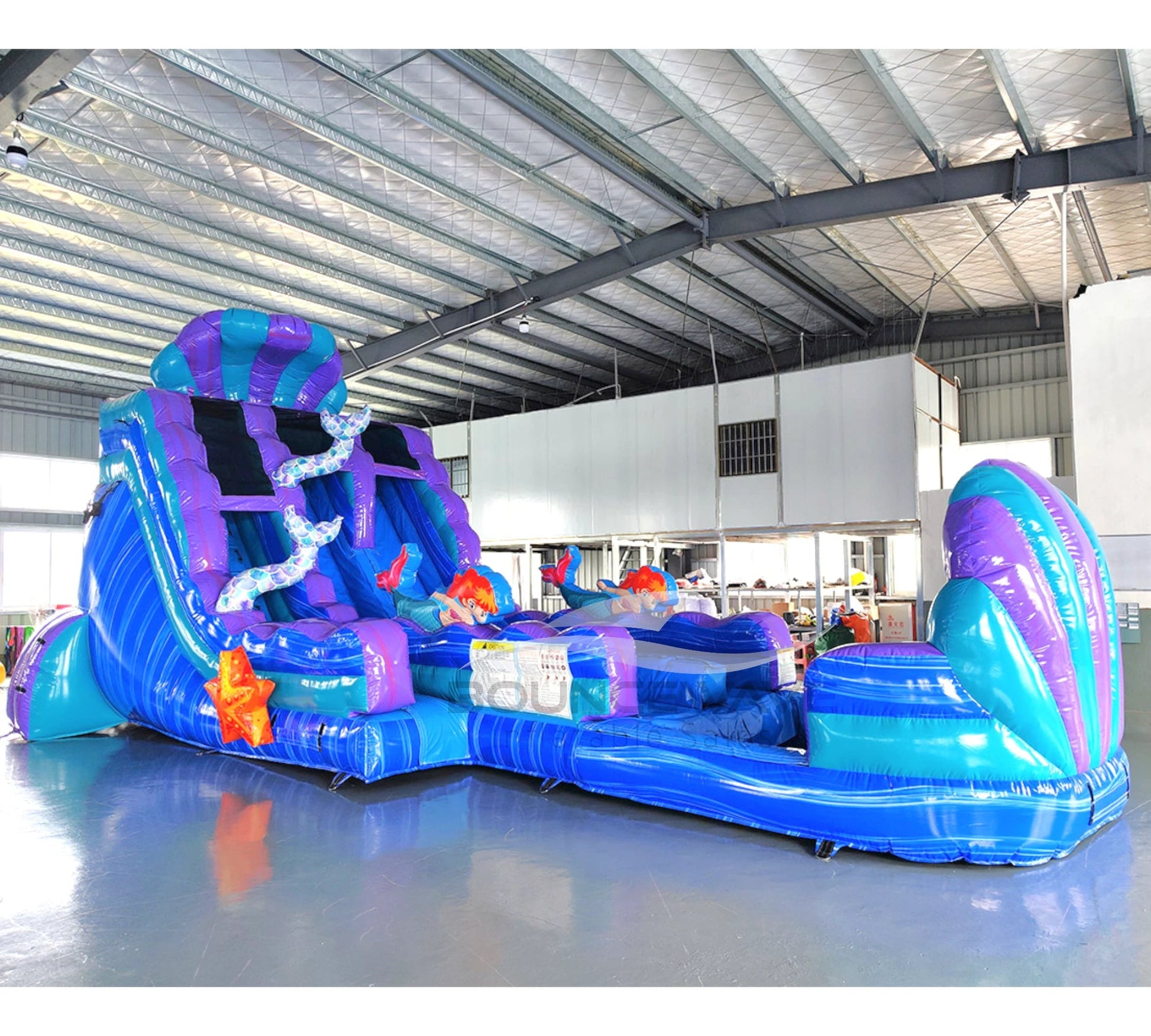 Front view of the 20ft Mermaid's Cove Hybrid DL inflatable water slide, showcasing its blue and purple sliding lanes, shell-shaped top, mermaid decorations, and spacious splash pool. The slide is set up in an indoor warehouse with other inflatables and equipment visible.