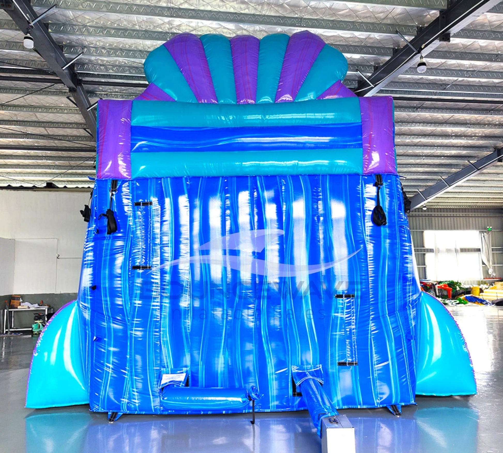 Side view of 20ft Mermaid's Cove Hybrid DL water slide showing vibrant blue, purple, and turquoise colors, shell-shaped design elements, and mermaid figures. The slide features both open and enclosed sliding sections, set up in an indoor facility with other inflatables visible in the background.