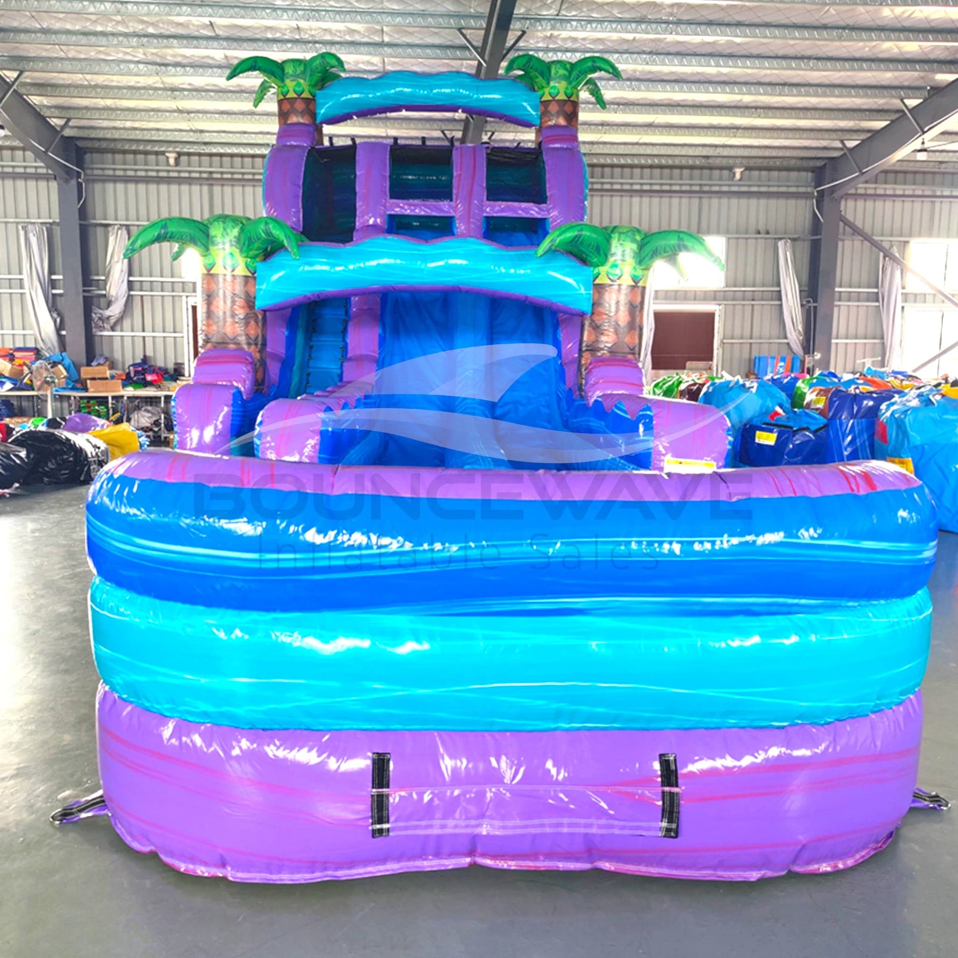 Front view of 20ft Purple Plunge Hybrid DL water slide featuring purple and blue colors, inflatable palm trees, and a large splash pool area. The slide is set up in an indoor facility with metal roofing and other inflatable products visible in the background.