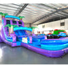 Front angle view of the 20ft Purple Plunge Hybrid DL inflatable water slide, showcasing its purple and blue sliding lanes, palm tree decorations, and spacious splash pool. The slide is set up in an indoor warehouse with other inflatables and equipment visible in the background.