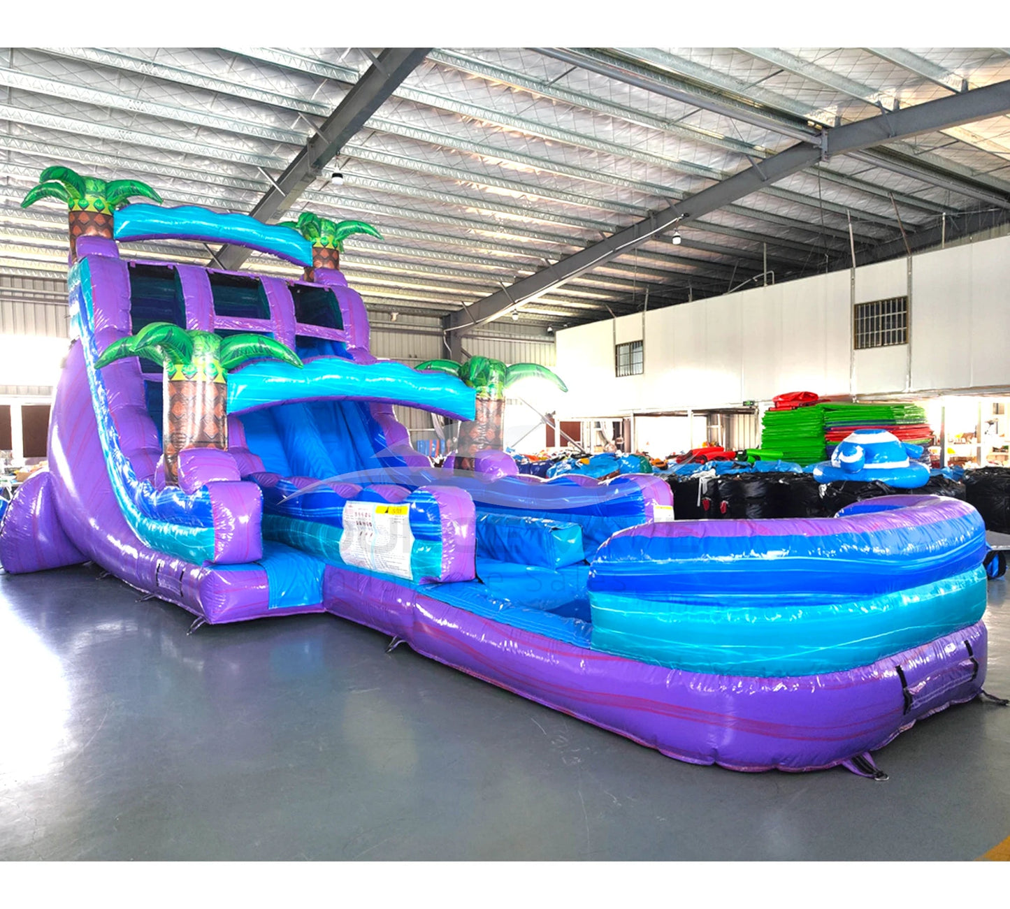 Front angle view of the 20ft Purple Plunge Hybrid DL inflatable water slide, showcasing its purple and blue sliding lanes, palm tree decorations, and spacious splash pool. The slide is set up in an indoor warehouse with other inflatables and equipment visible in the background.