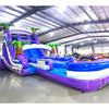 Front angle view of the 20ft Purple Thunder Hybrid DL inflatable water slide, showcasing its purple and blue sliding lanes, palm tree decorations, and spacious splash pool. The slide is set up in an indoor warehouse with other inflatables and equipment visible in the background.