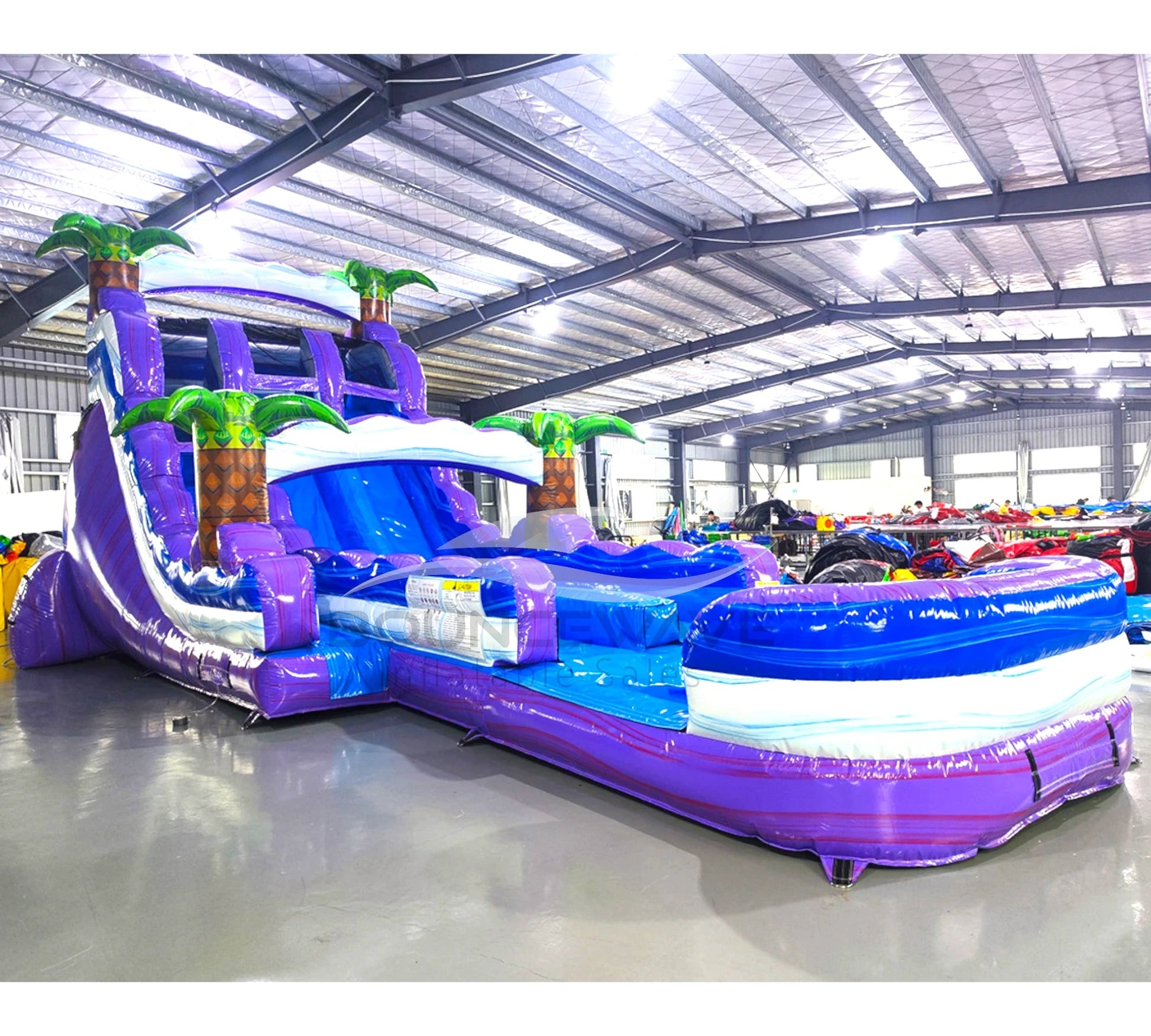 Front angle view of the 20ft Purple Thunder Hybrid DL inflatable water slide, showcasing its purple and blue sliding lanes, palm tree decorations, and spacious splash pool. The slide is set up in an indoor warehouse with other inflatables and equipment visible in the background.