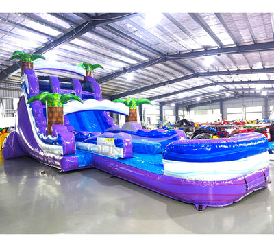 Front angle view of the 20ft Purple Thunder Hybrid DL inflatable water slide, showcasing its purple and blue sliding lanes, palm tree decorations, and spacious splash pool. The slide is set up in an indoor warehouse with other inflatables and equipment visible in the background.