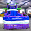 Front view of 20ft Purple Thunder Hybrid DL water slide featuring purple and blue colors, inflatable palm trees, and a large splash pool area with "BOUNCEWAVE" branding. The slide is set up in an indoor facility with metal roofing and other inflatable products visible in the background.