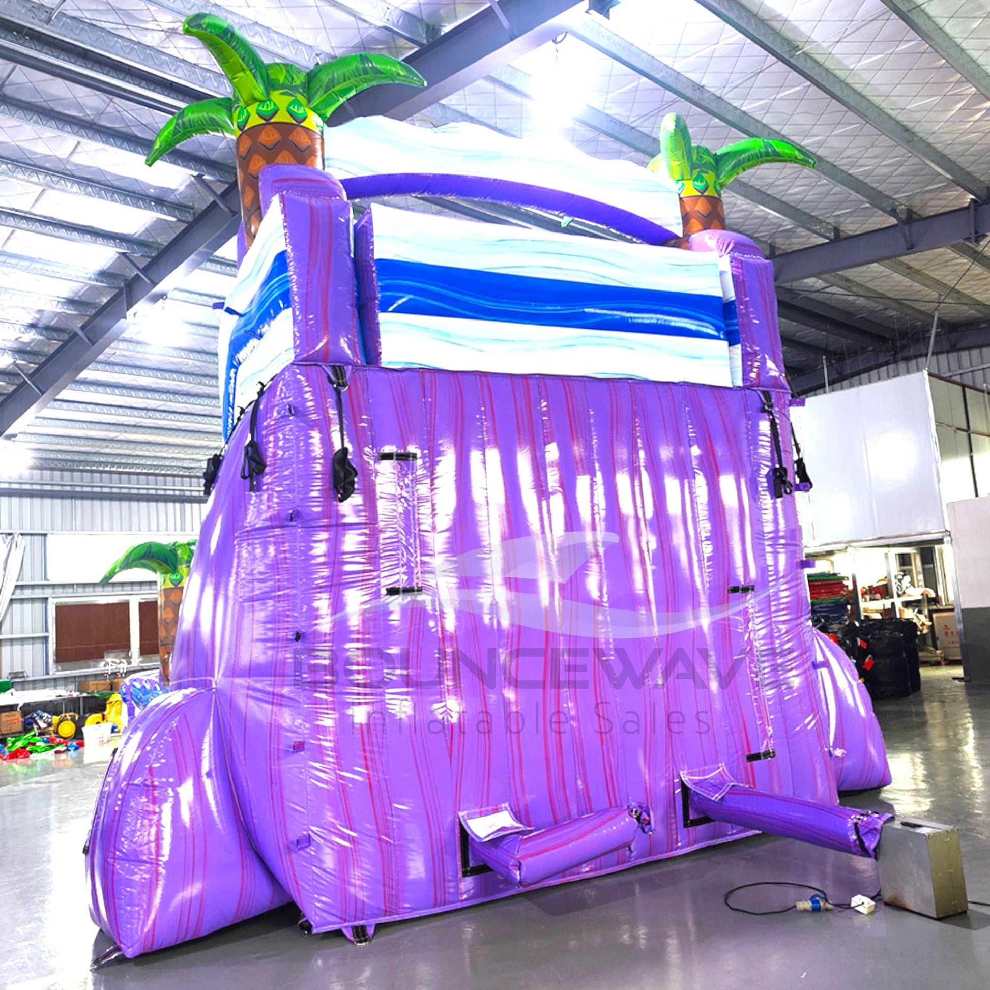 Large inflatable water slide with tropical theme, featuring vibrant purple and blue colors, palm tree decorations, and a spacious splash pool area. The slide combines open and enclosed sections, set up in an indoor warehouse with metal roofing and other equipment visible in the background.