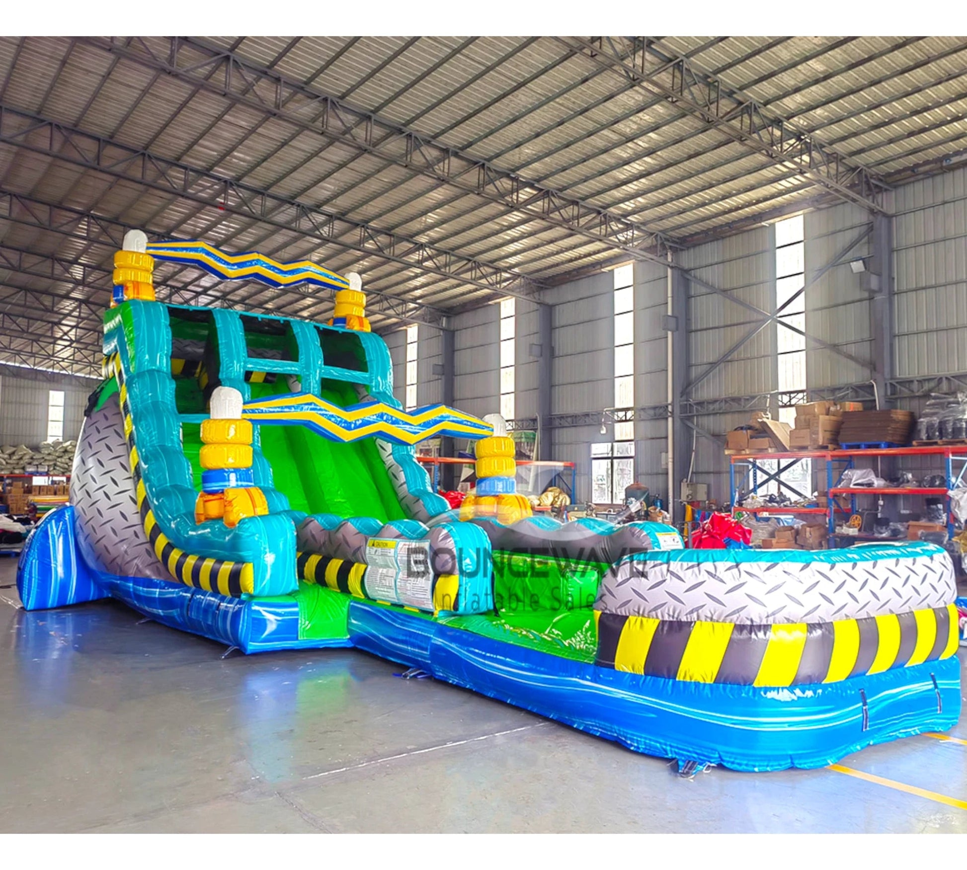 Side angle view of 20ft Radioactive Run Hybrid DL water slide featuring bright green and blue colors, caution stripe patterns, and a large splash pool area. The slide is set up in an indoor facility with metal roofing, disco balls, and other inflatables visible in the background.