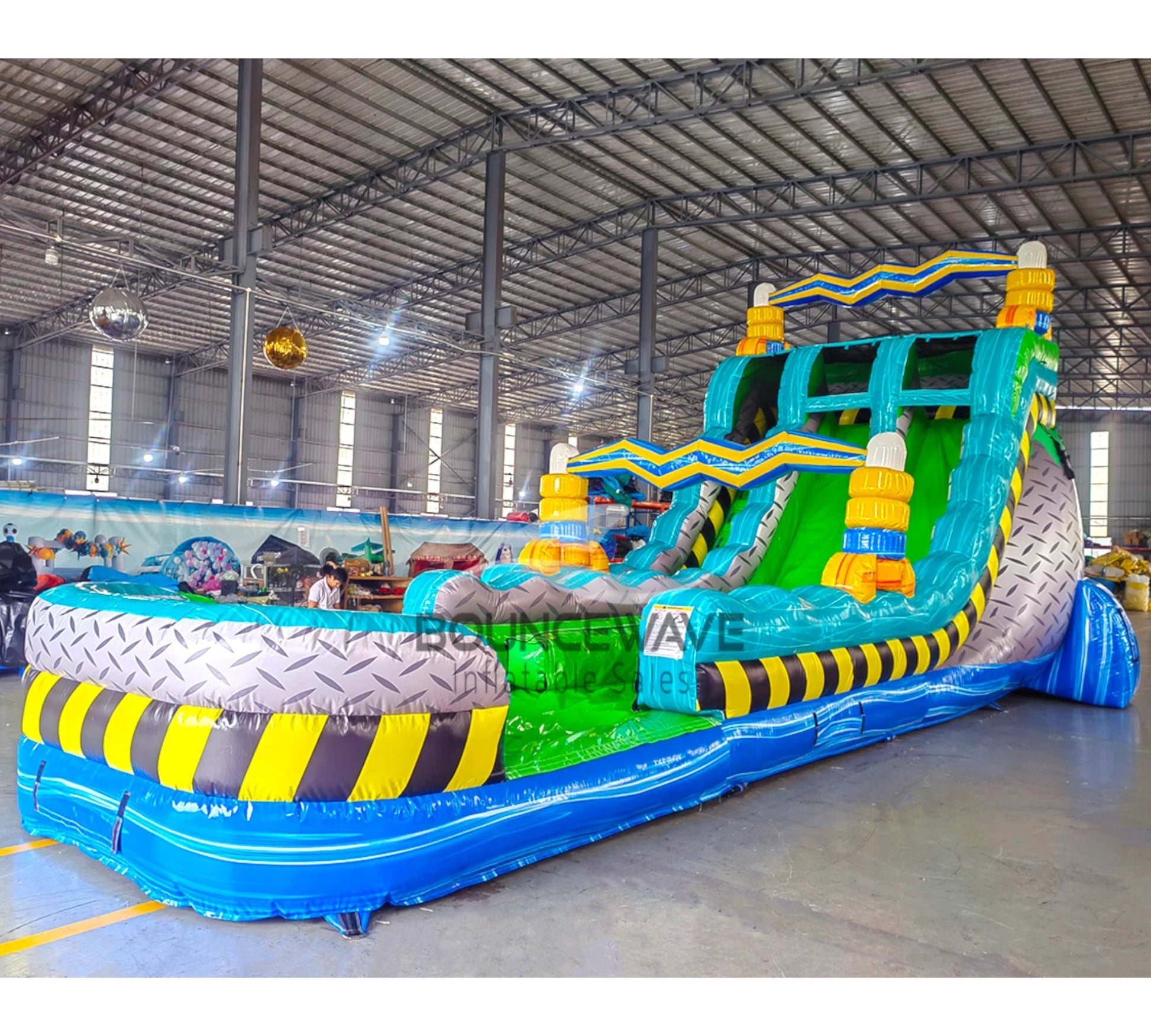 Front view of the 20ft Radioactive Run Hybrid DL inflatable water slide, showcasing its bright green sliding lanes, blue accents, caution stripe decorations, and spacious splash pool. The slide is set up in an indoor warehouse with other inflatables and equipment visible.